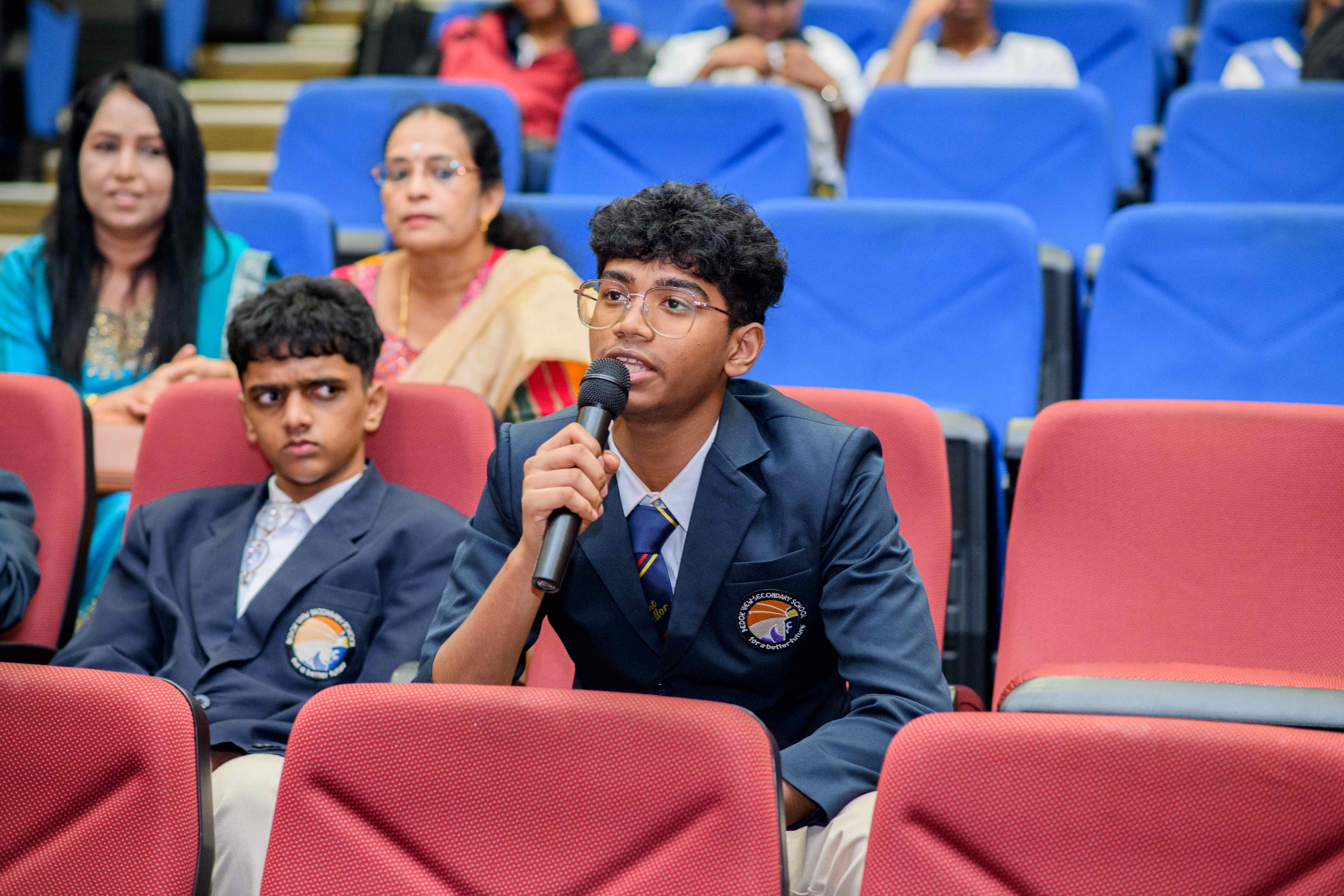 Student in navy blazer speaks into a microphone in an auditorium; other students and adults sit behind.