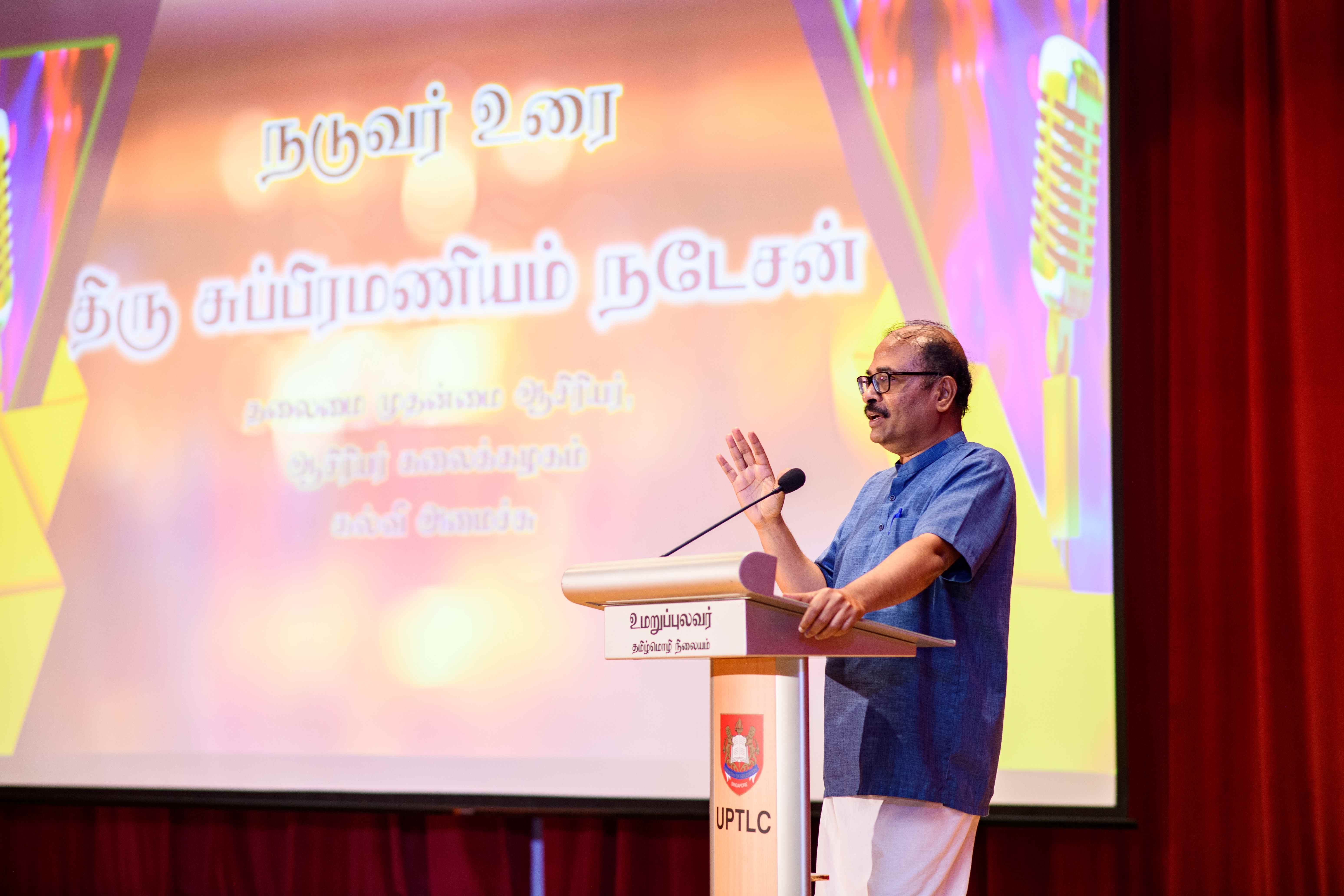 Man in blue shirt speaks at podium on stage, hand raised, with Tamil text projected behind.