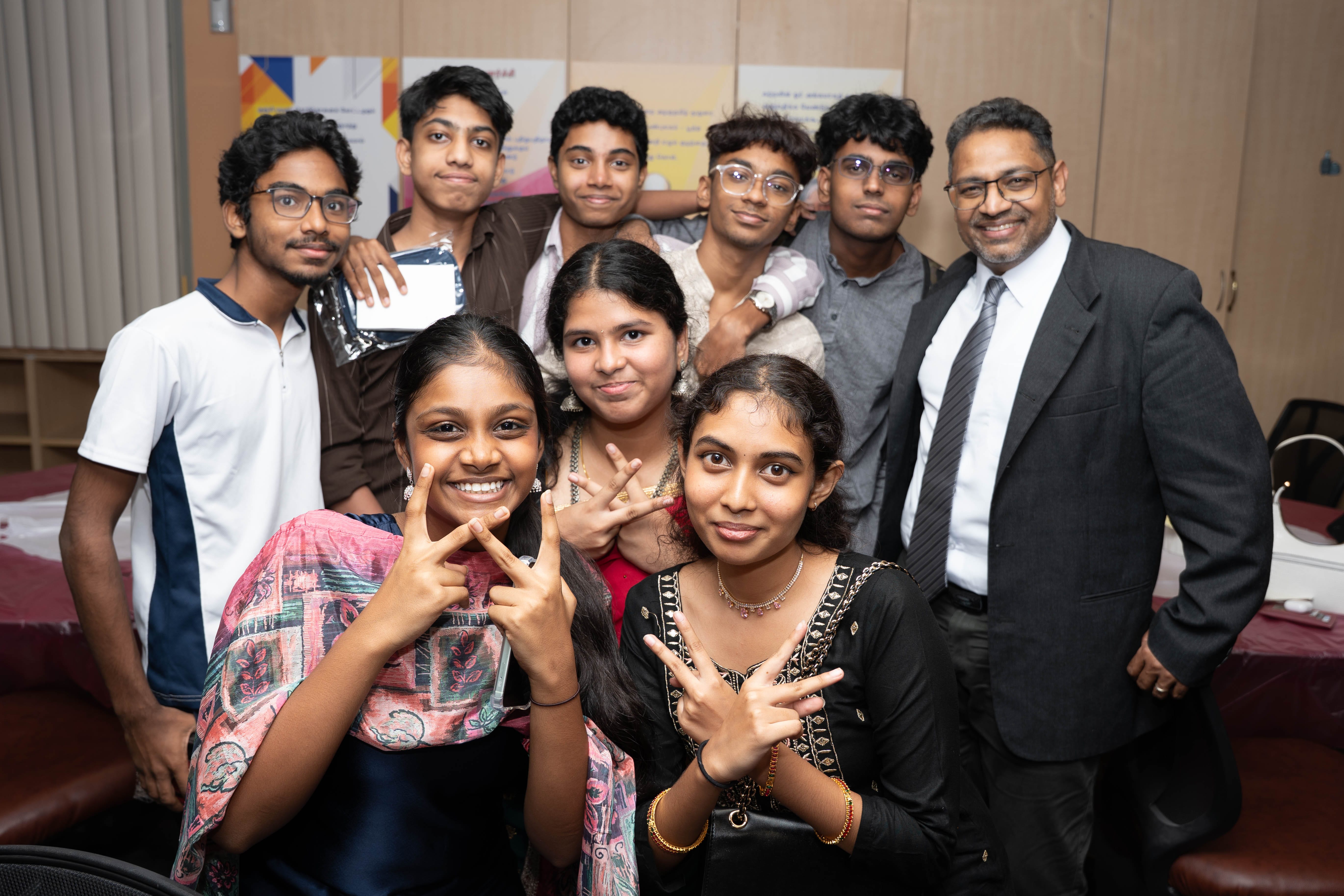 Group of nine smiling people posing indoors; three in front make peace signs, one man in suit at right.
