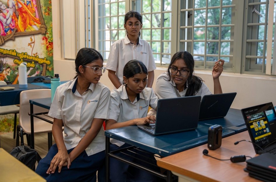 Four schoolgirls in uniforms work at desks, focused on a laptop in a classroom with large windows.