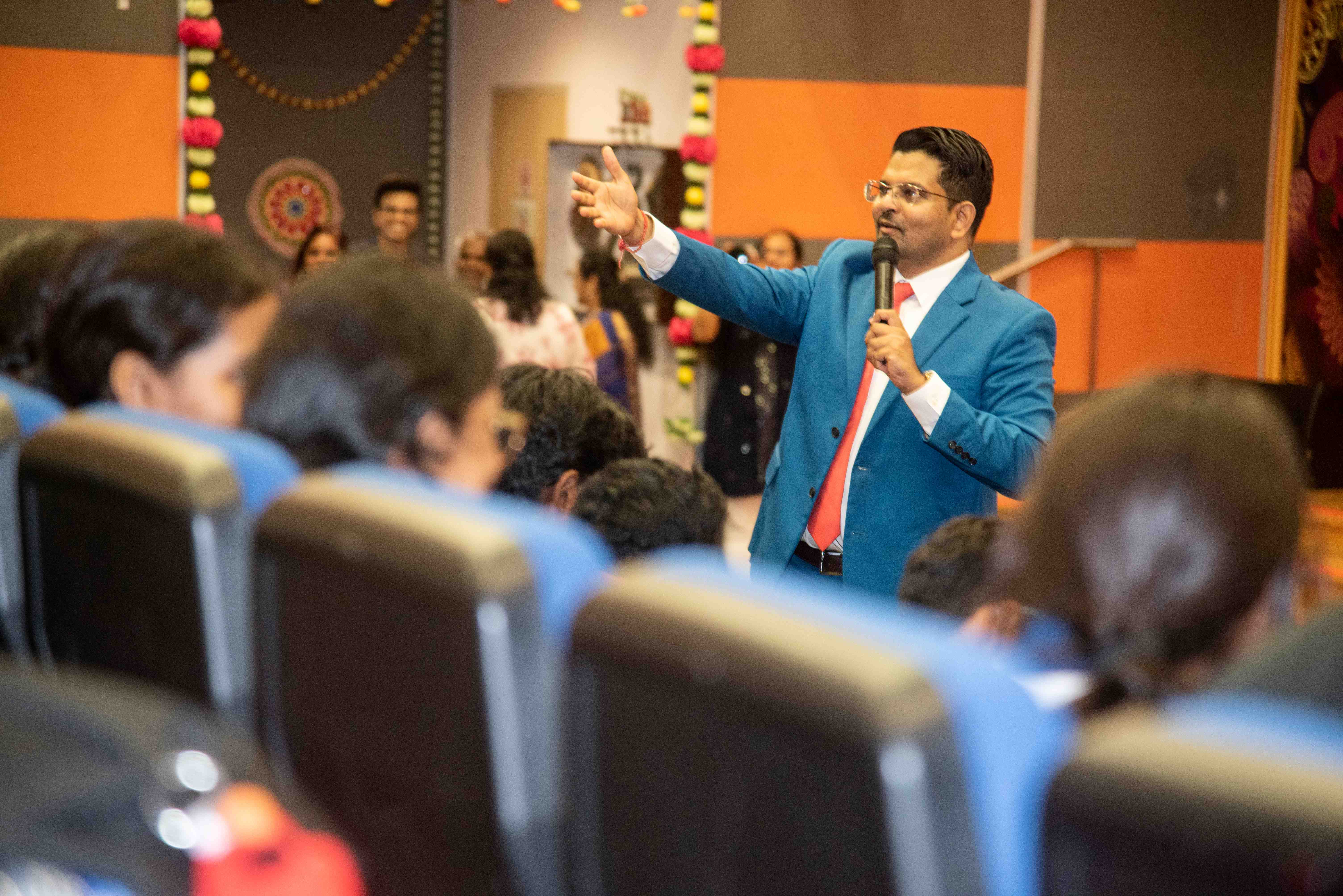 Man in blue suit holds a microphone and gestures while speaking to seated audience in an auditorium.