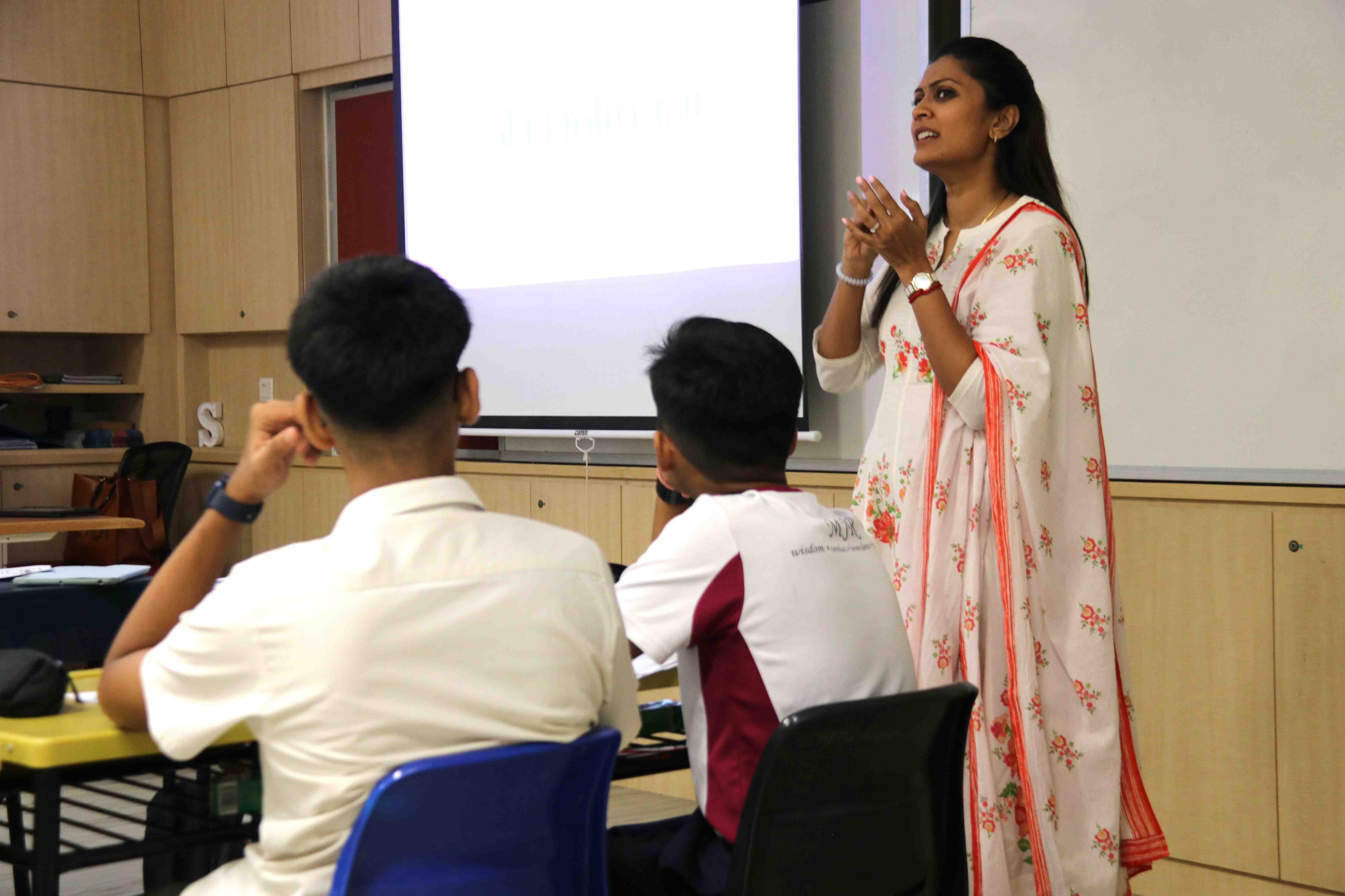 Woman in floral dress speaks beside a projection screen as two seated students face her in a classroom.