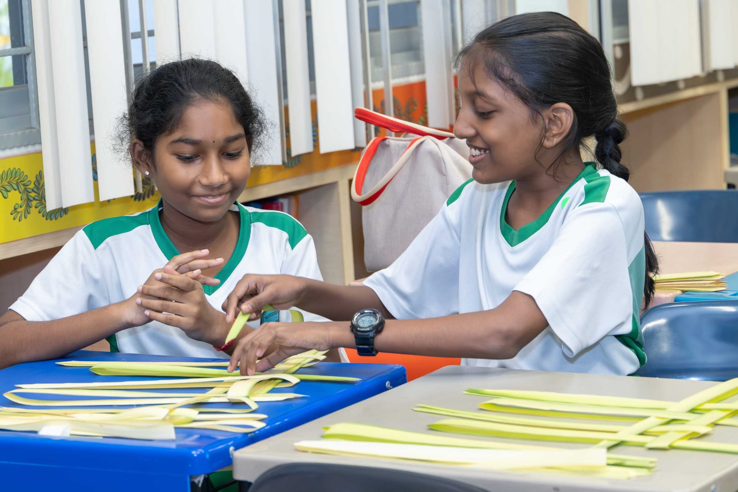 Two schoolgirls in white and green uniforms smile while weaving pale strips on classroom tables.