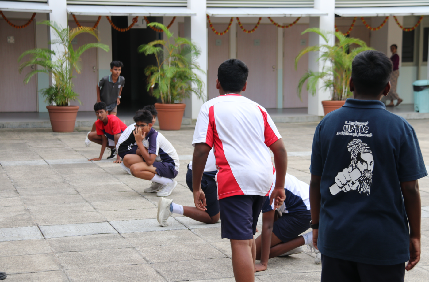 Students in sports uniforms play a game in a school courtyard with potted palms and doors behind.