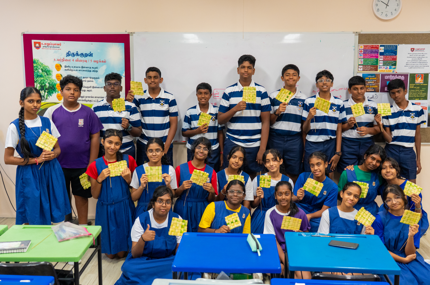 Students in school uniforms pose in a classroom holding woven palm-leaf squares, with a whiteboard behind.