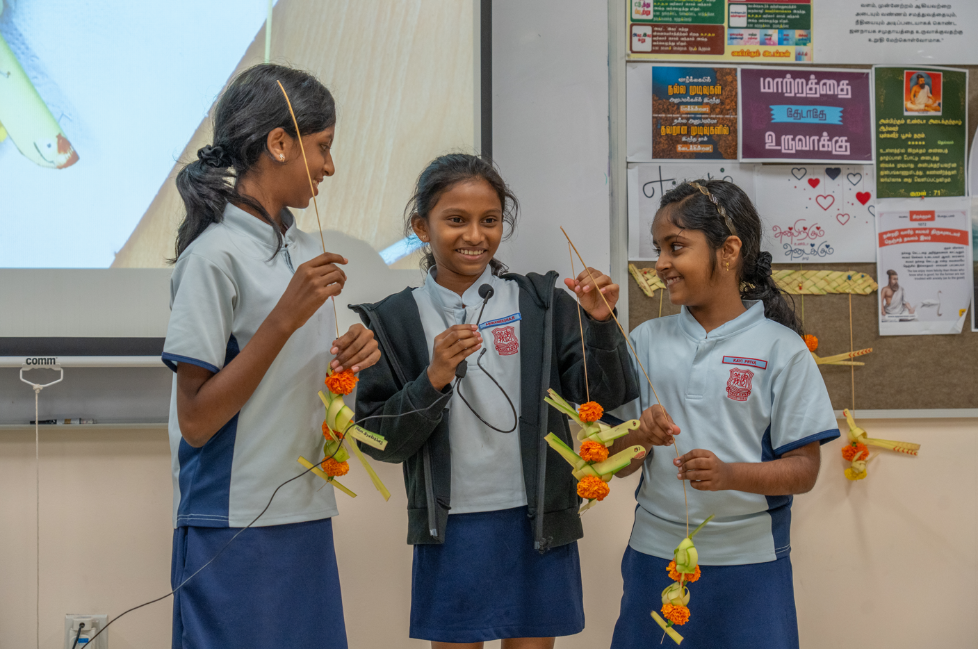 Three schoolgirls in uniforms smile and hold hanging palm-leaf and marigold decorations in a classroom.