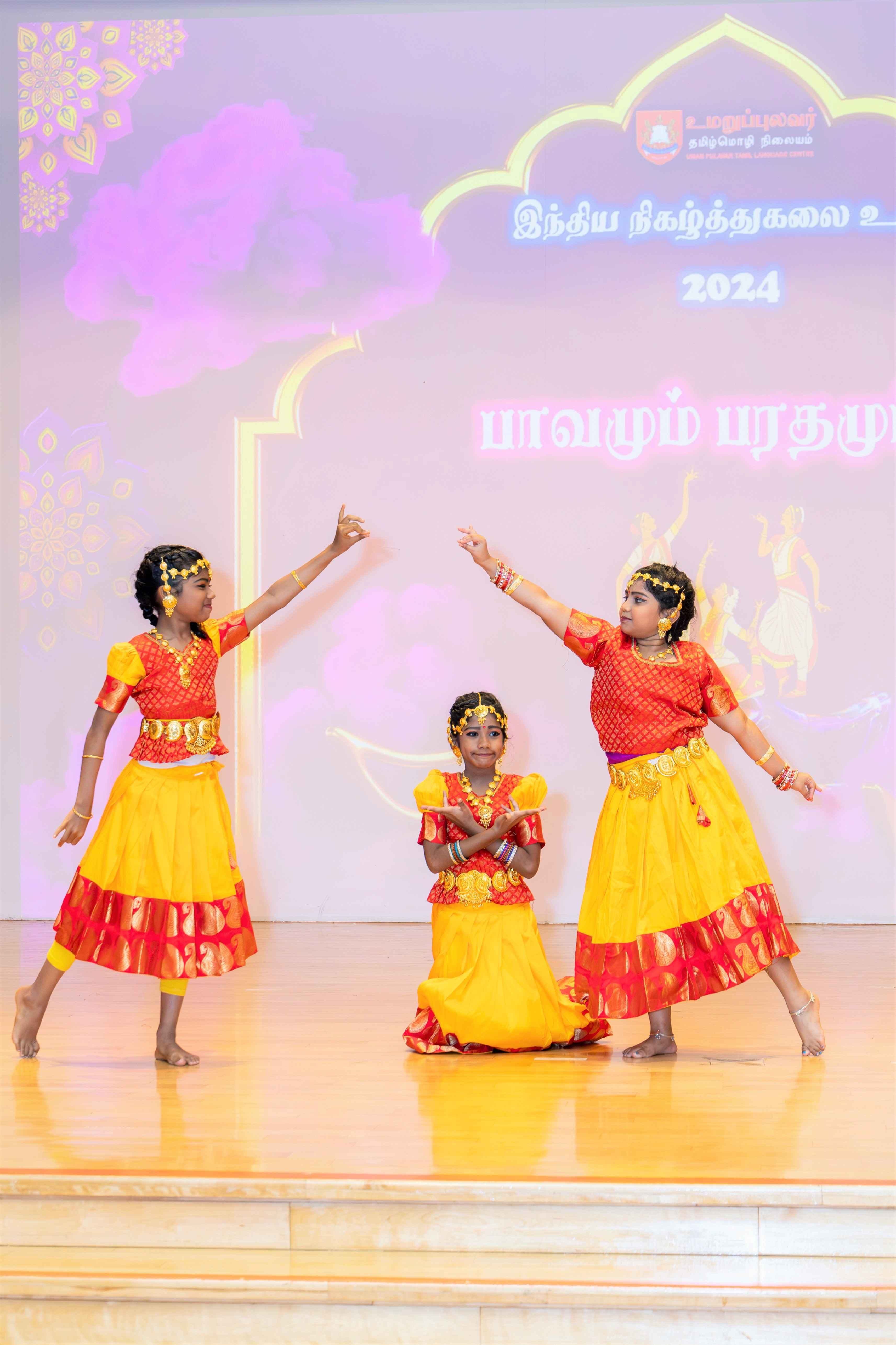 Three girls in red and yellow dance costumes pose on stage, two standing with raised arms, one kneeling.