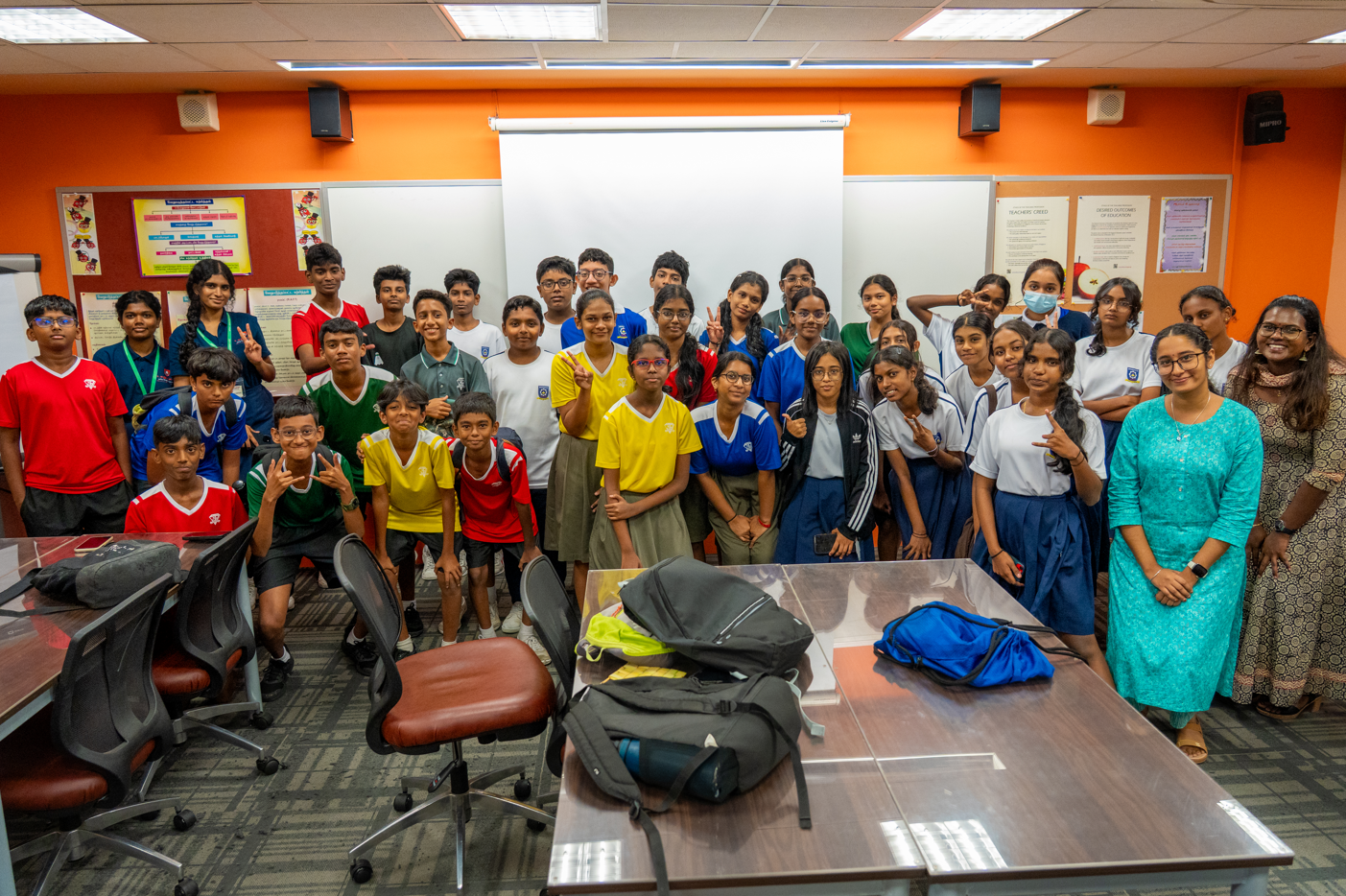 Group of students and two adults posing in a classroom, with chairs and backpacks on tables in front