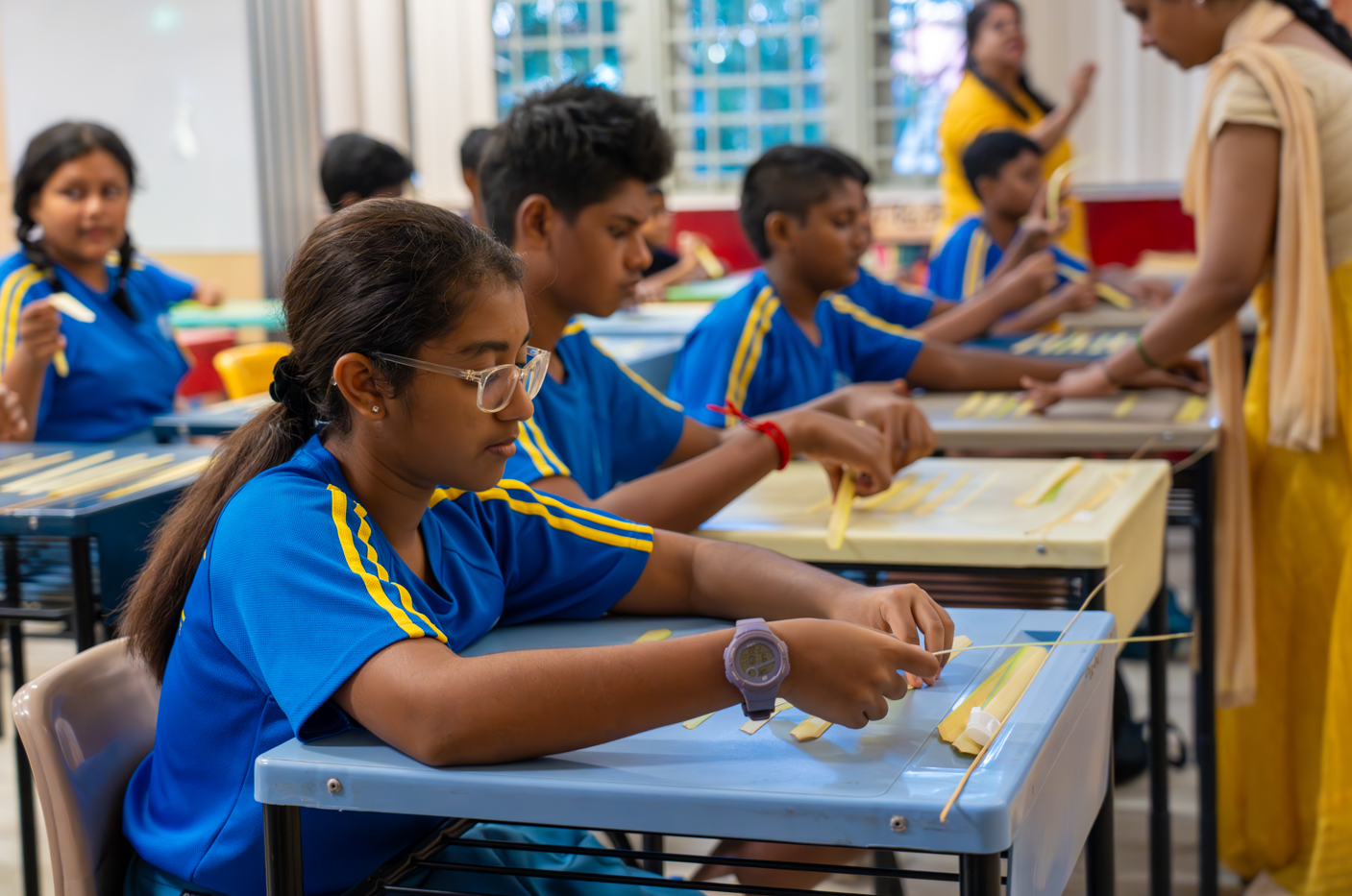 Students in blue uniforms work with thin sticks at desks while a teacher in yellow assists nearby.