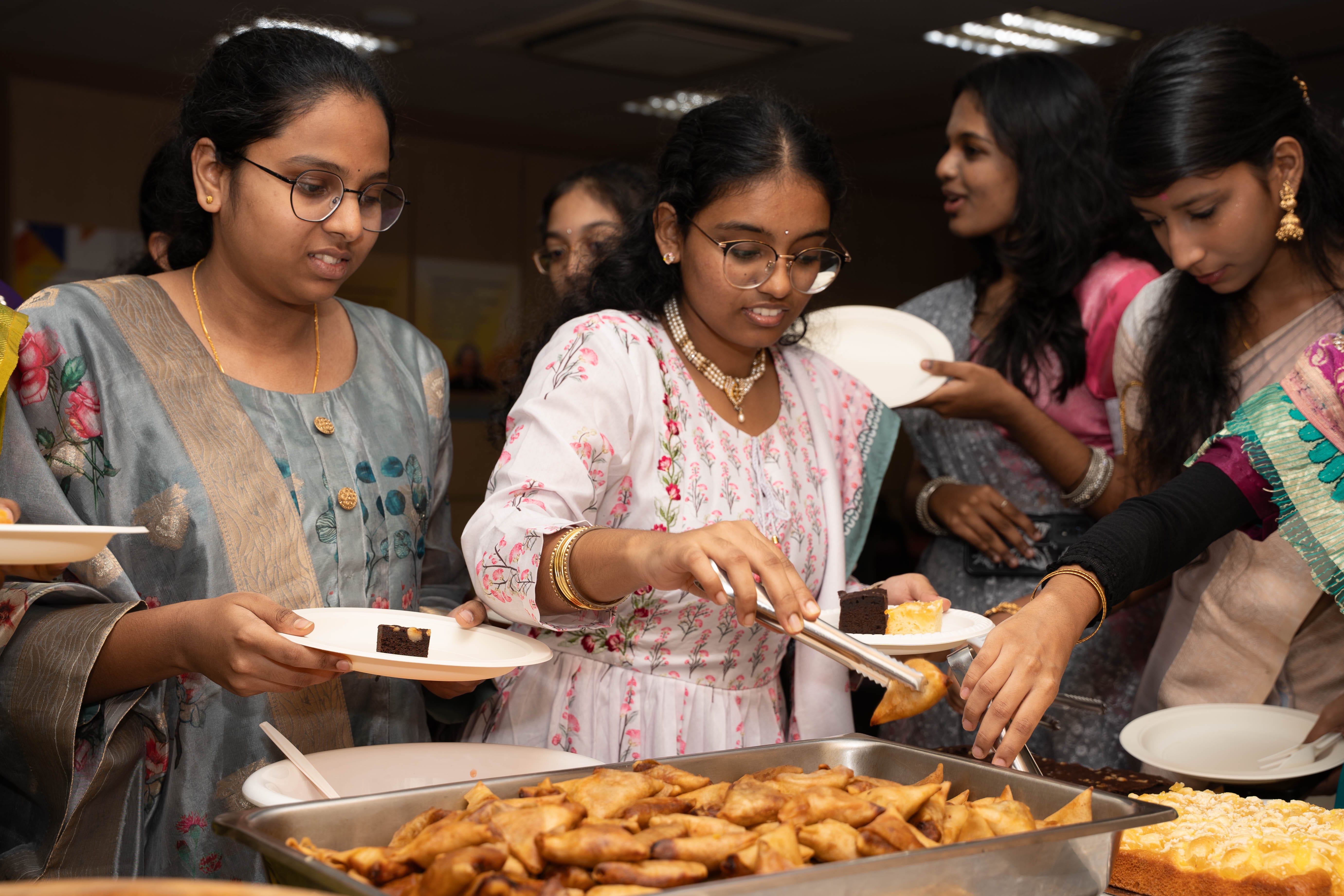 Women in traditional outfits serve pastries and cake onto plates at a buffet table indoors.