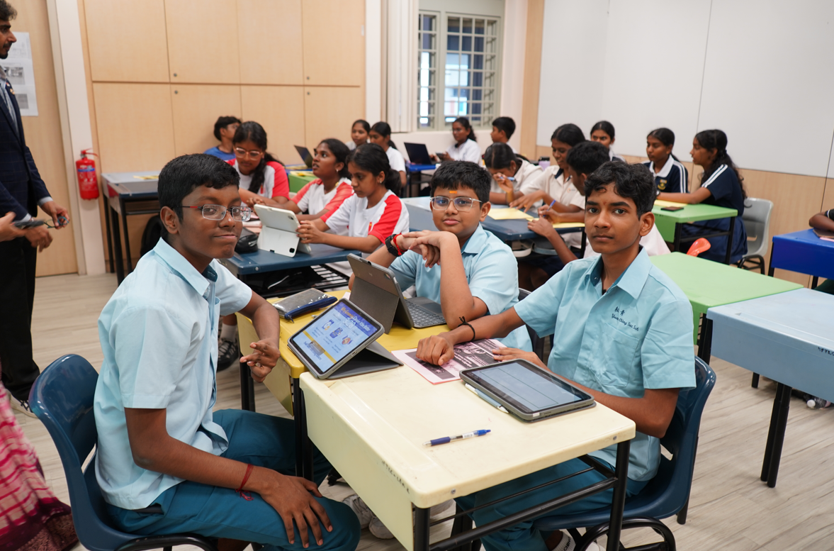 Students in a classroom sit at desks with tablets and notebooks, three boys in light blue uniforms in front.