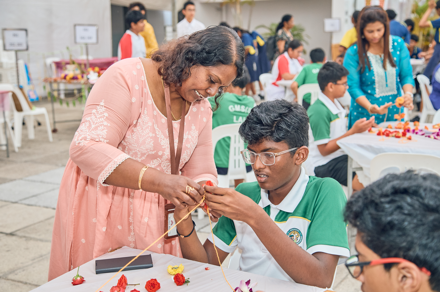 Woman in pink leans over, helping a seated student in green shirt craft with string and flowers.