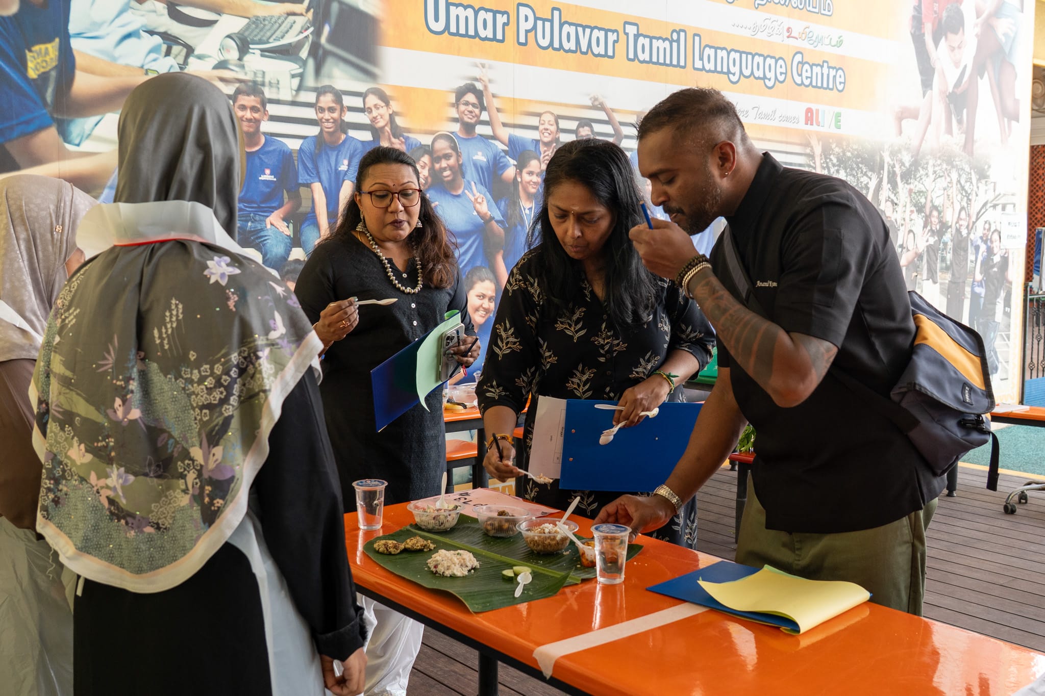 Group tasting food from small bowls on a banana leaf at a table under a Tamil language centre banner.