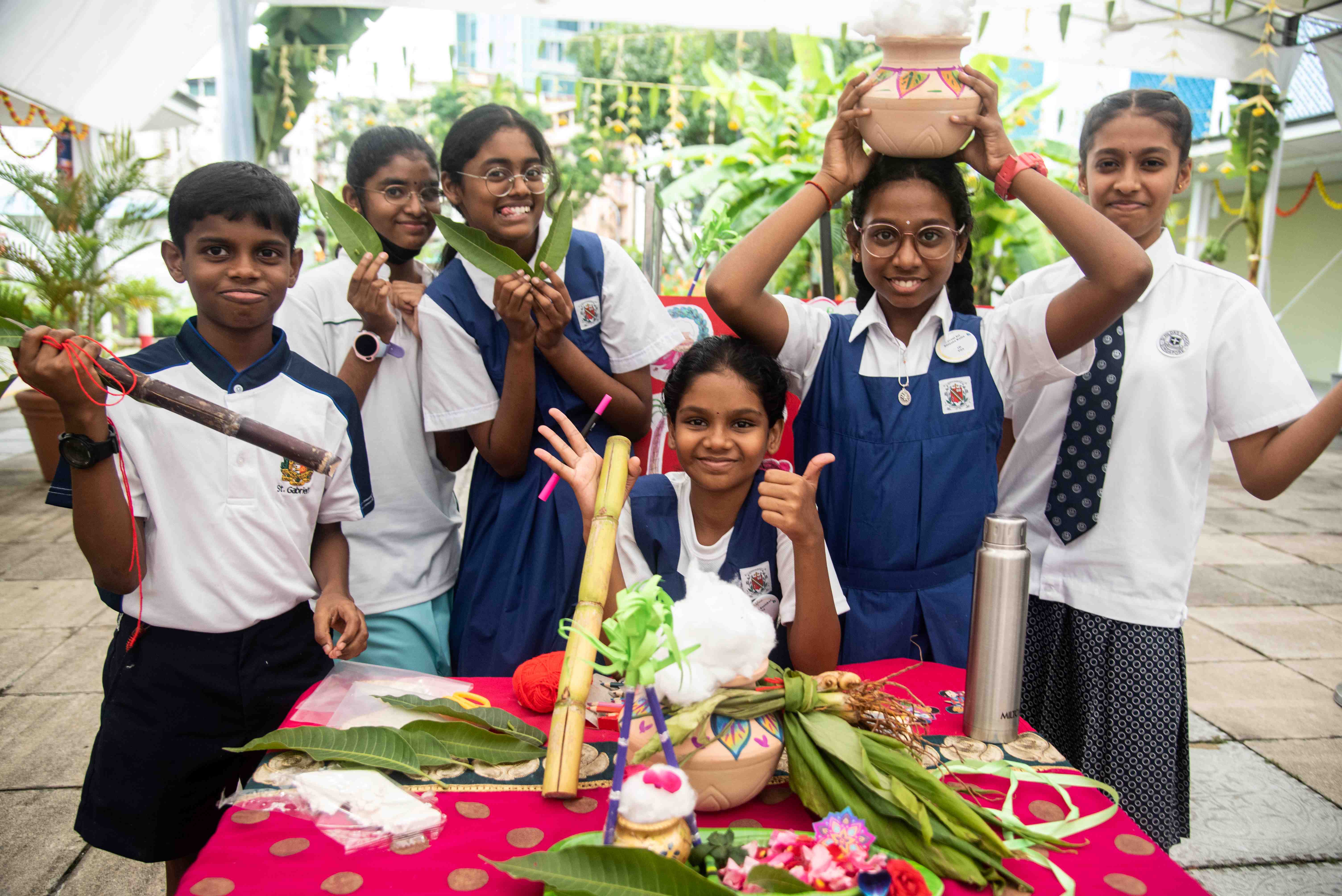 Six schoolchildren in uniforms smile at a table with leaves, sticks, crafts, and a girl holding a pot on her head.