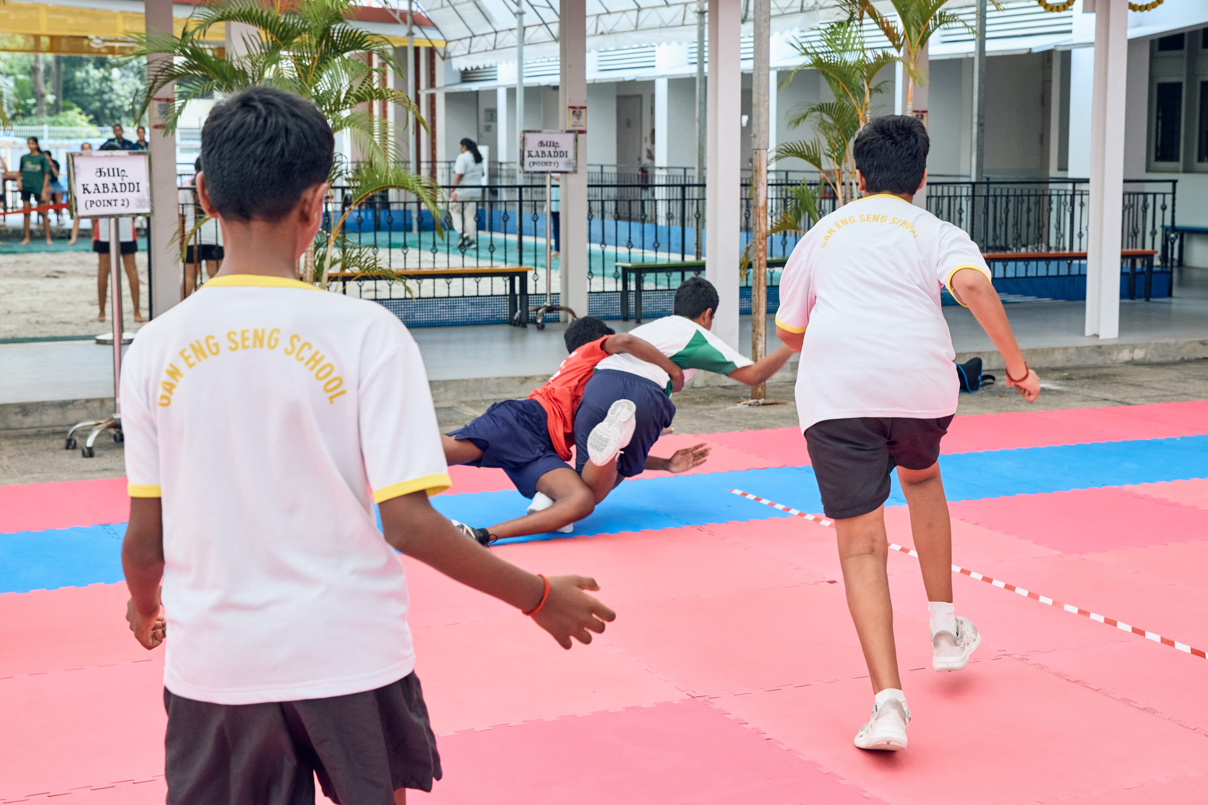 Boys play kabaddi on red mats as one tackles another, with teammates running in a covered courtyard.
