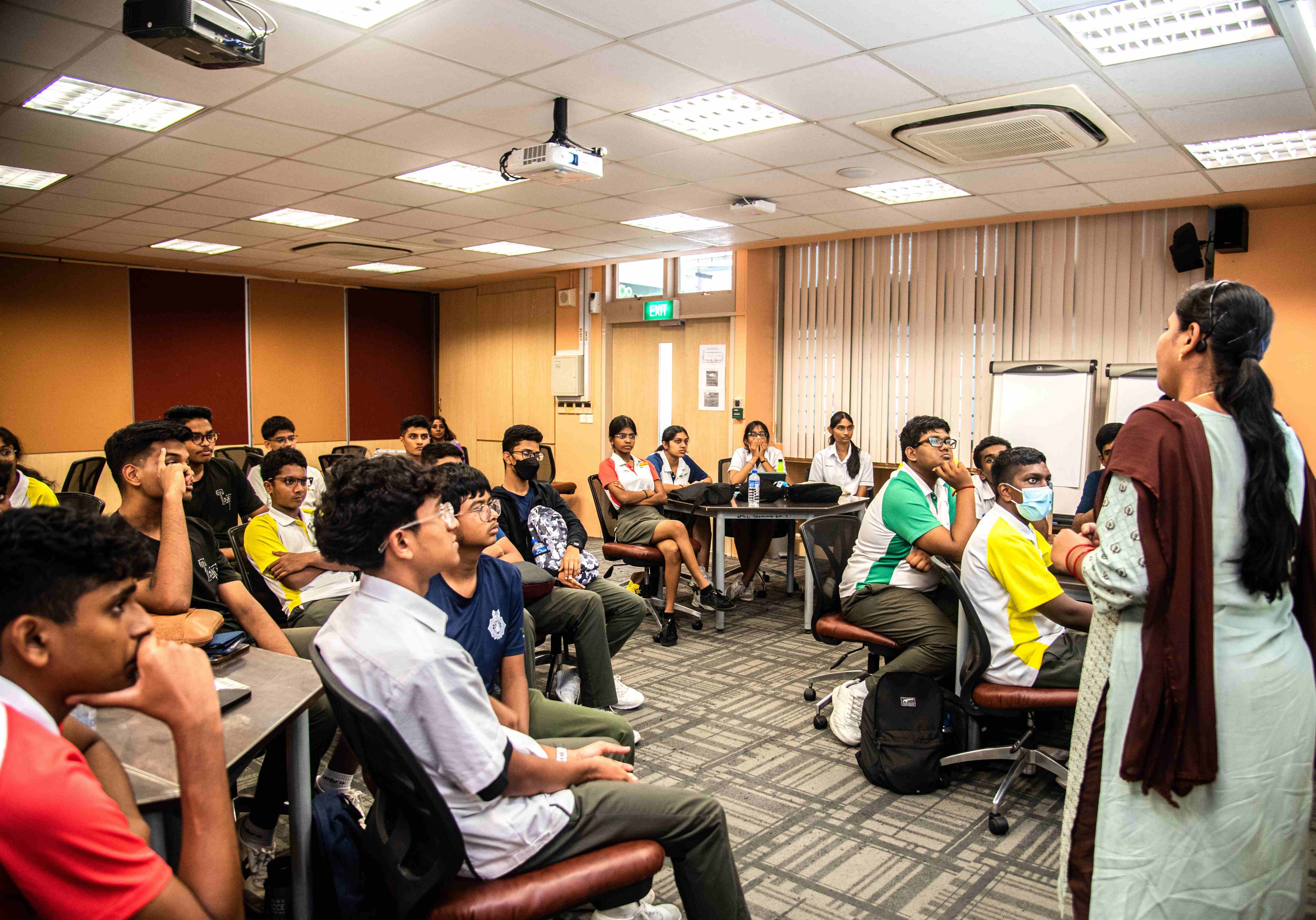 Teacher stands at front of classroom speaking to seated students, with ceiling projector and whiteboards behind.