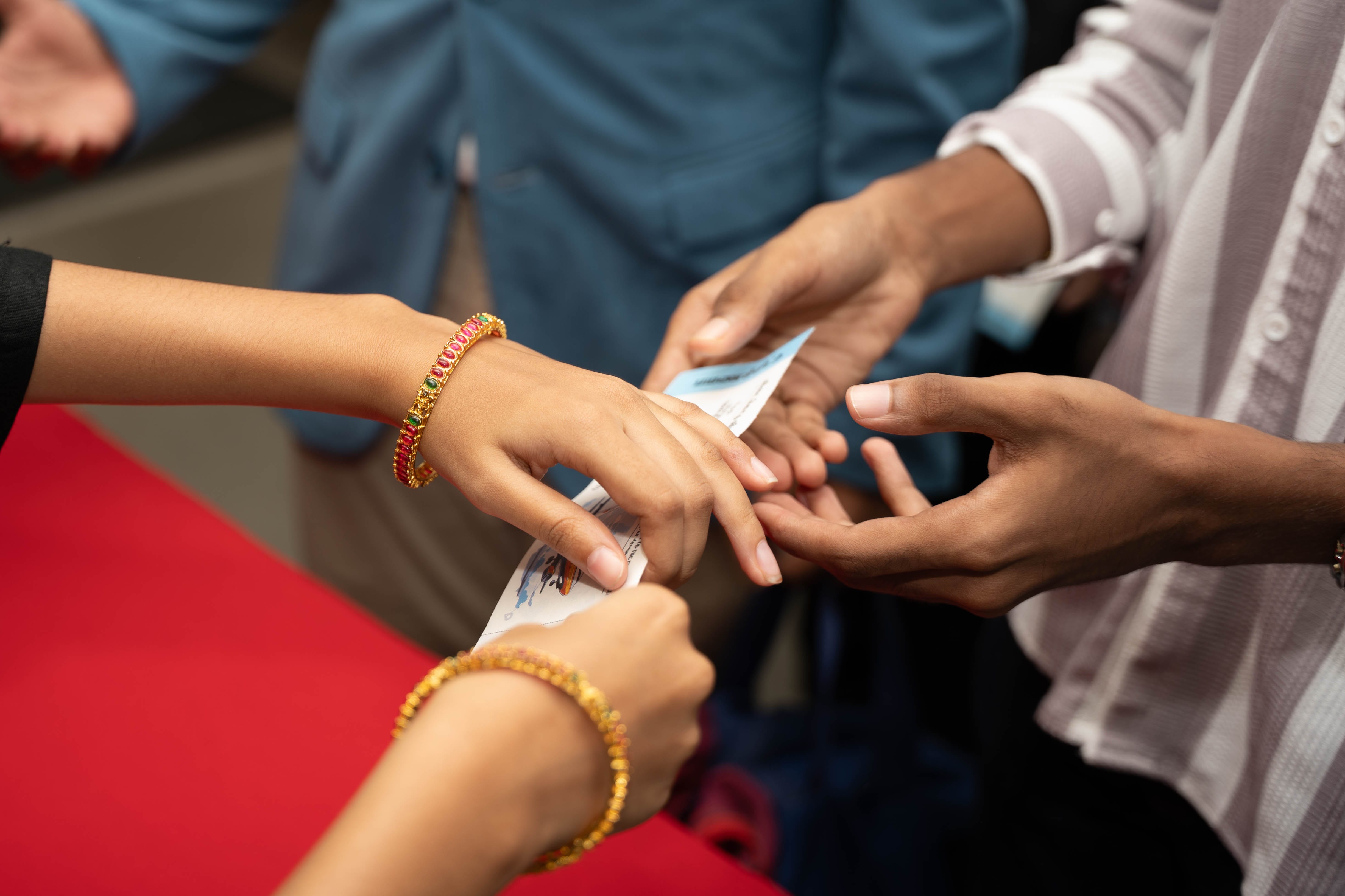 Close-up of hands exchanging paper tickets; two wrists wear gold and red bangles.