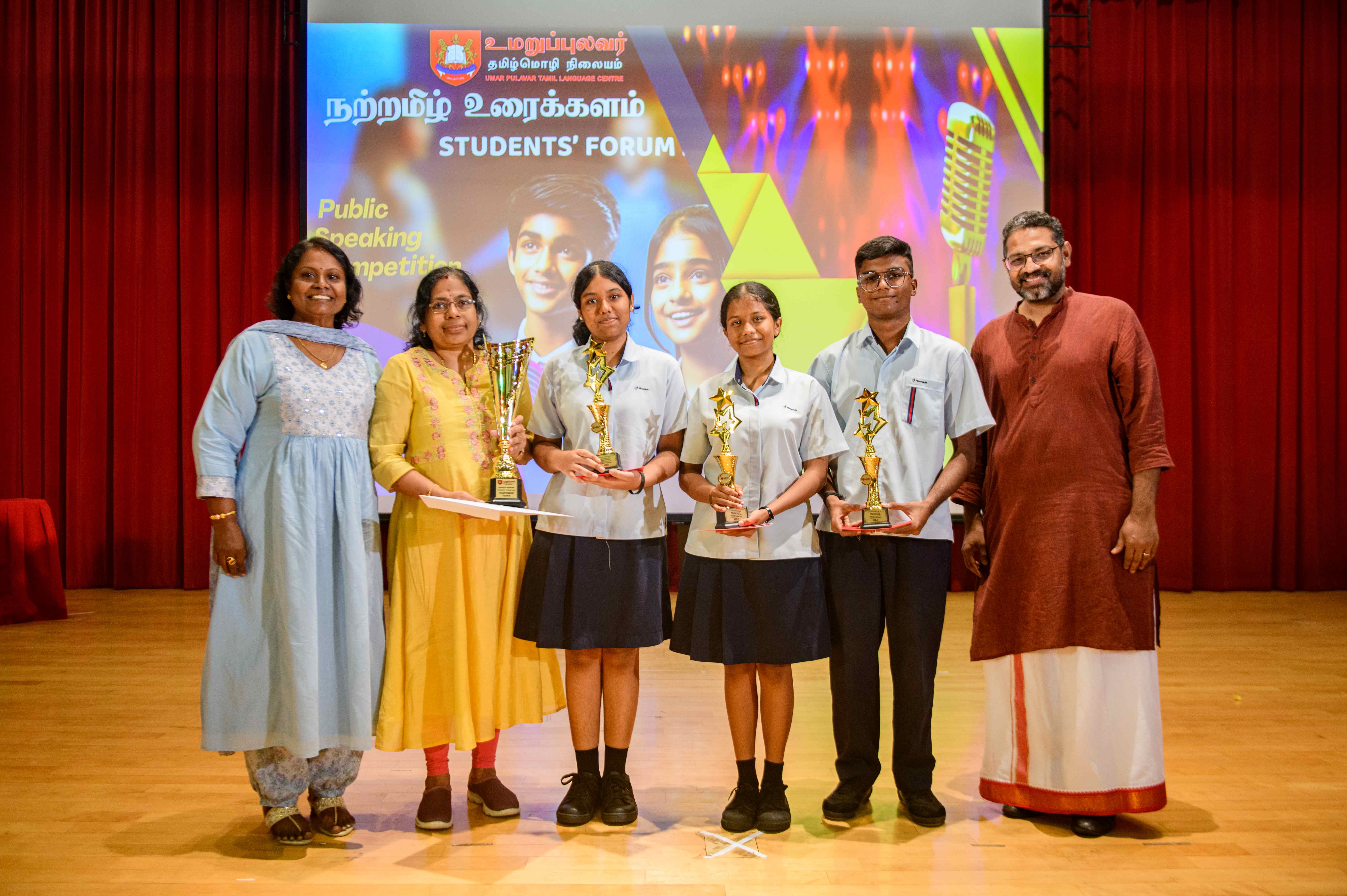 Group on stage holding trophies in front of Students' Forum public speaking competition banner and red curtains