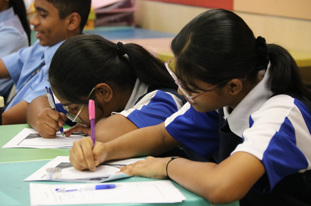 Two schoolgirls in blue uniforms lean over worksheets, writing with pens at a classroom table.