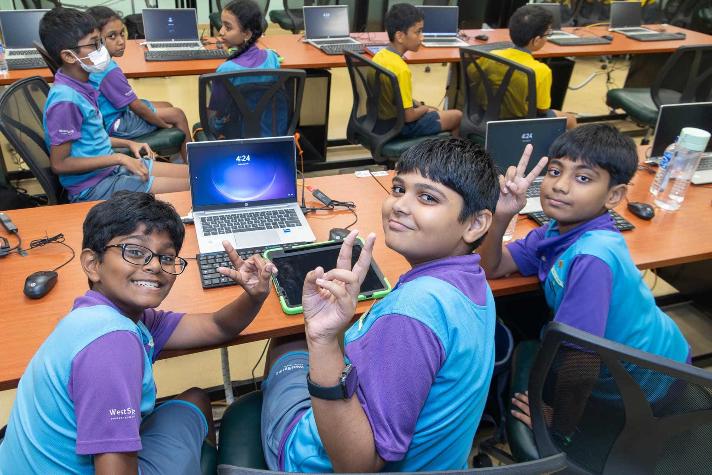 Schoolchildren in blue and purple uniforms sit at computers; three boys smile and flash peace signs.