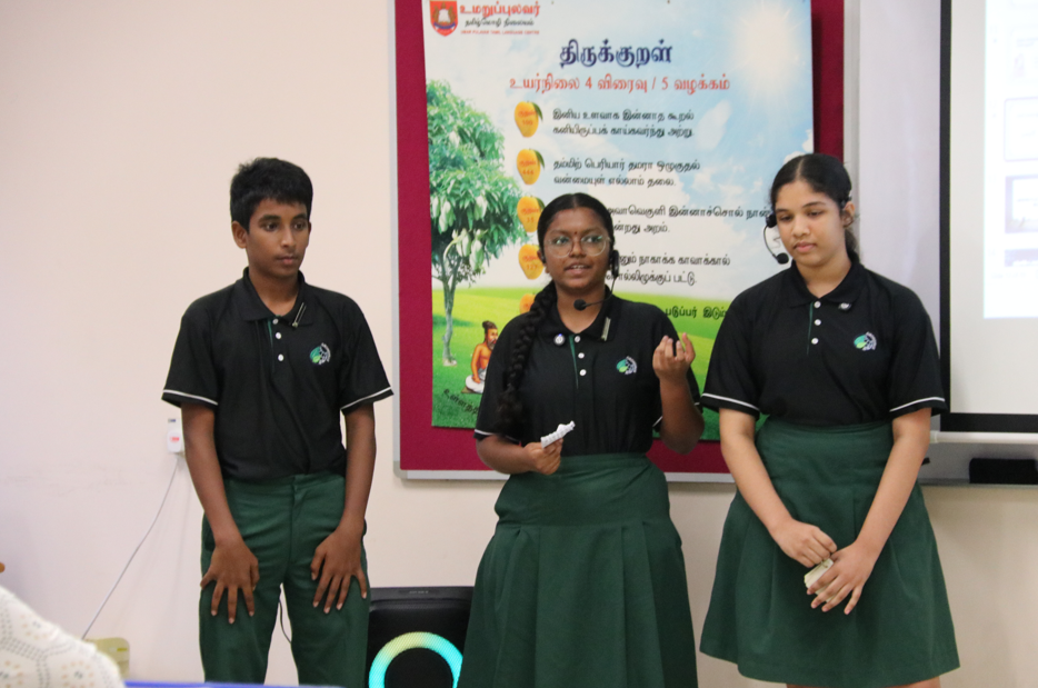 Three students in black shirts and green uniforms give a presentation in front of a Tamil poster and projector.