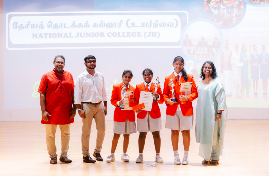 Six people on stage; three students in red blazers hold certificates and trophies under a college banner.