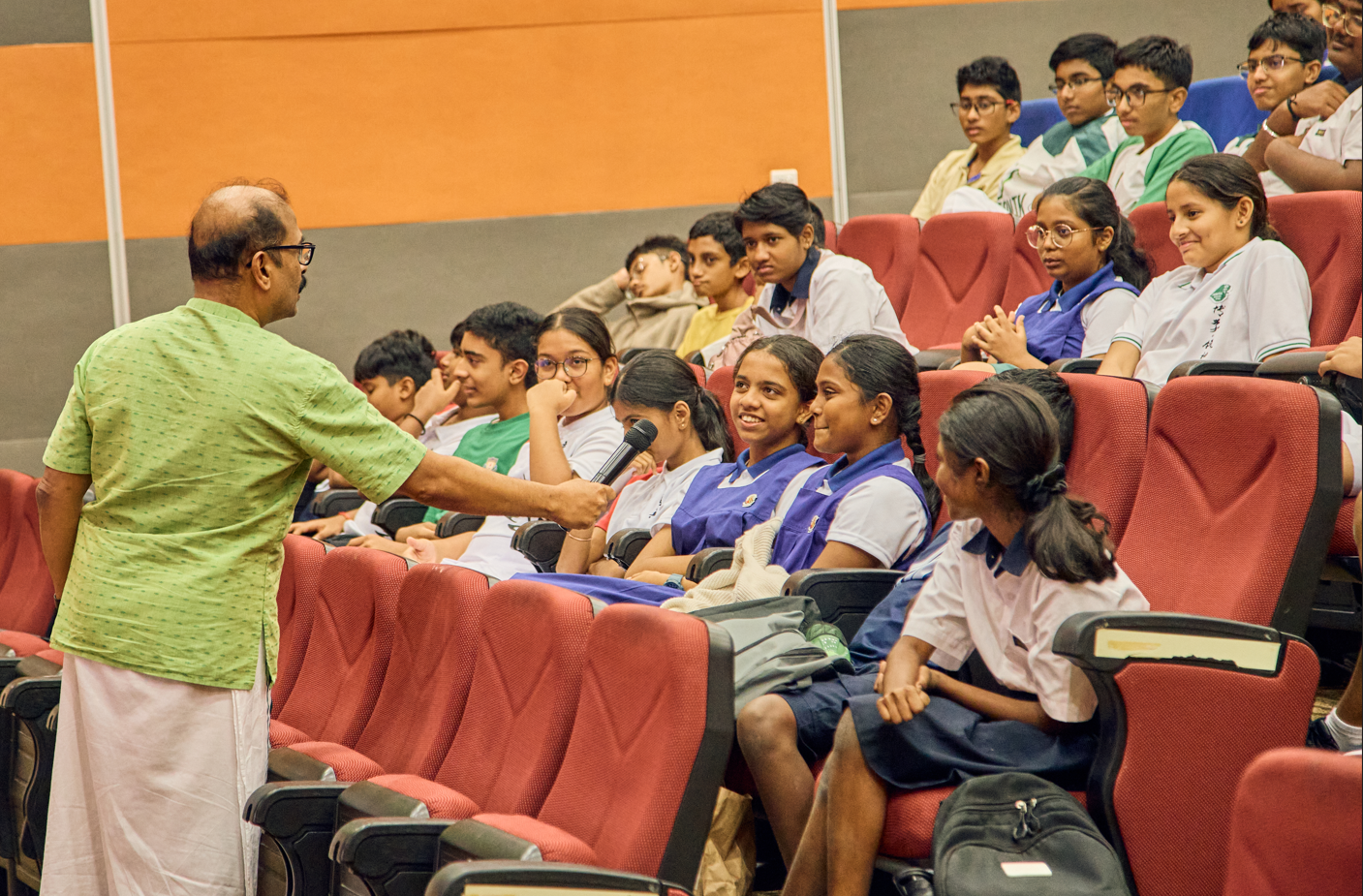 Man with microphone interviews seated students in school uniforms in a lecture hall with red seats.