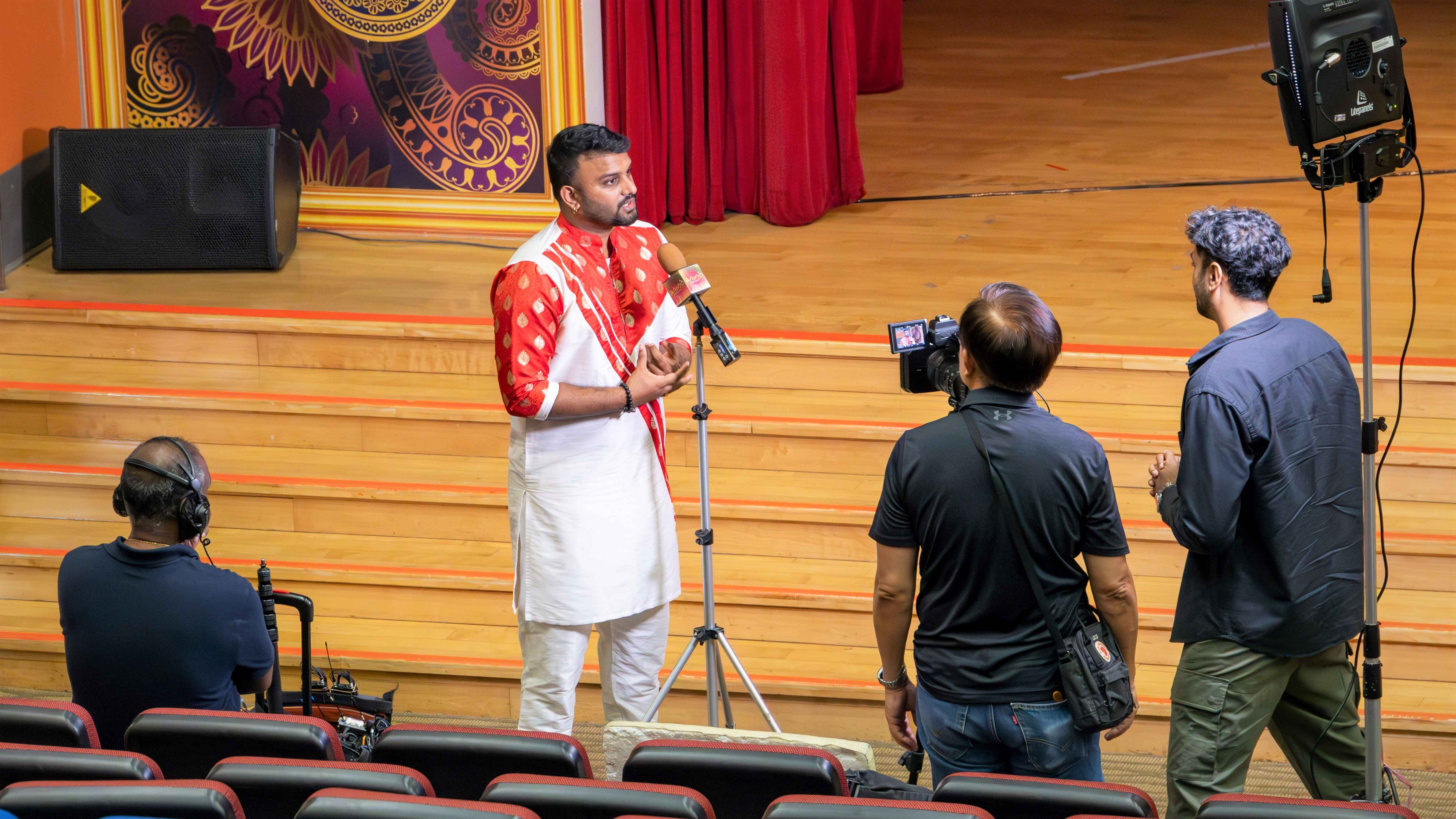 Man in red and white outfit speaks at a mic on stage while a crew films him with camera and light.