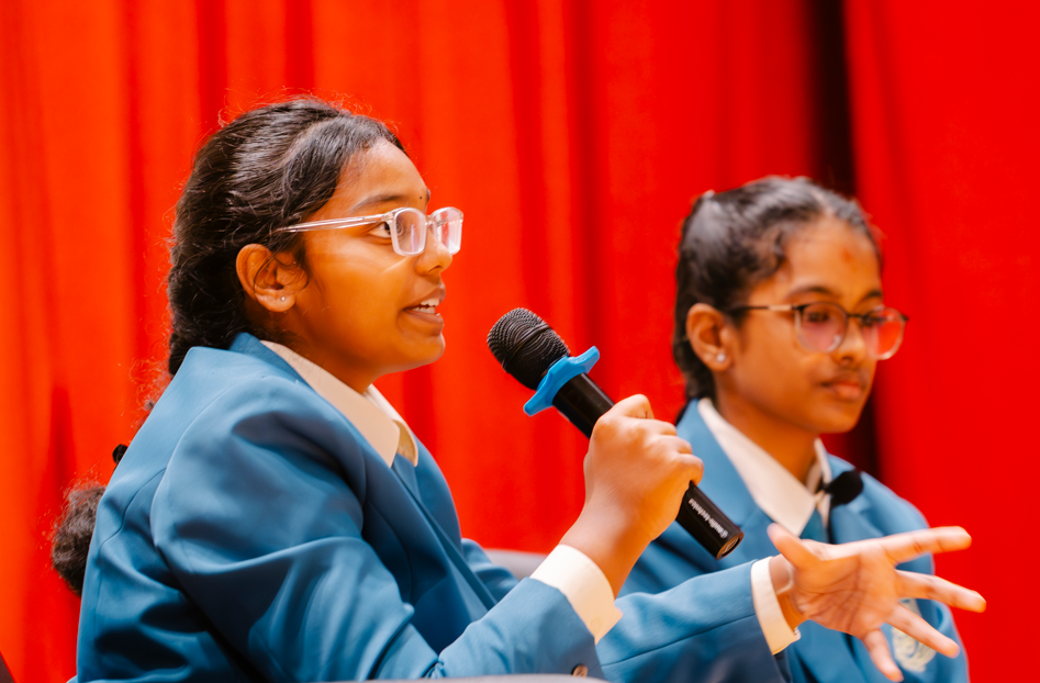 Student in blue blazer speaks into a handheld microphone onstage, with another student seated beside her.