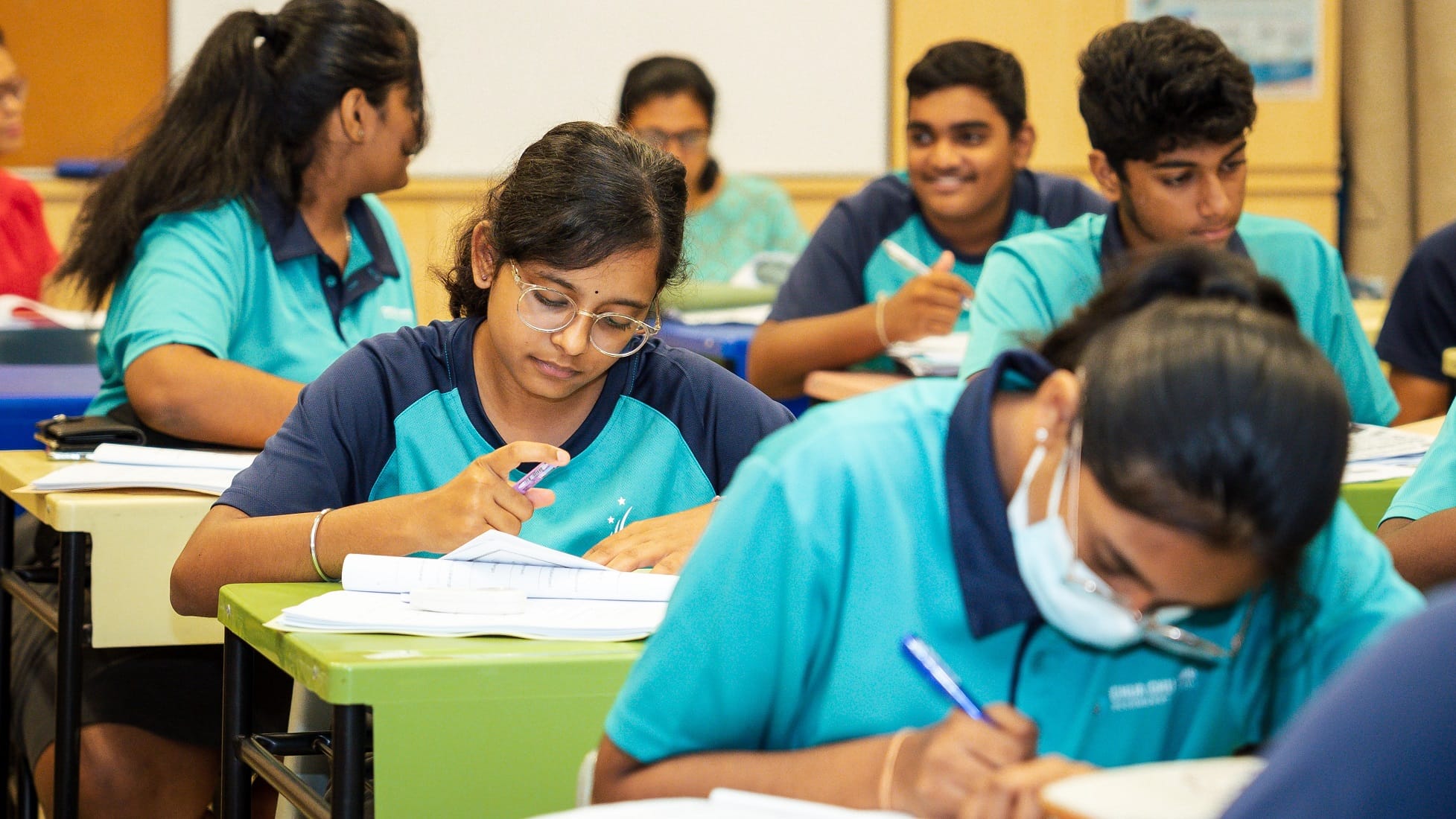 Students in teal uniforms sit at desks, writing in notebooks; one girl with glasses is centered.