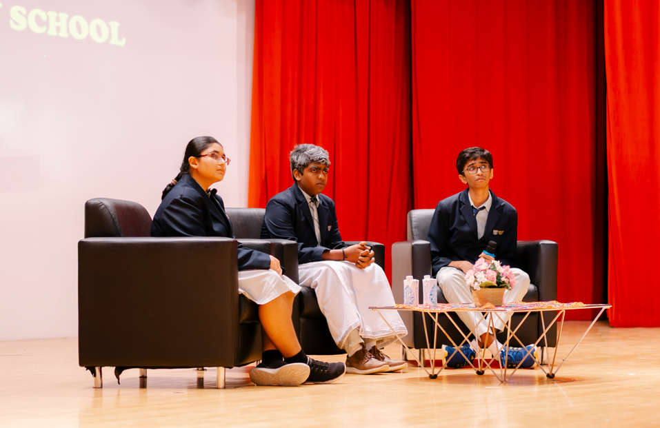 Three students in blazers sit on black chairs onstage by a table with flowers, against red curtains.