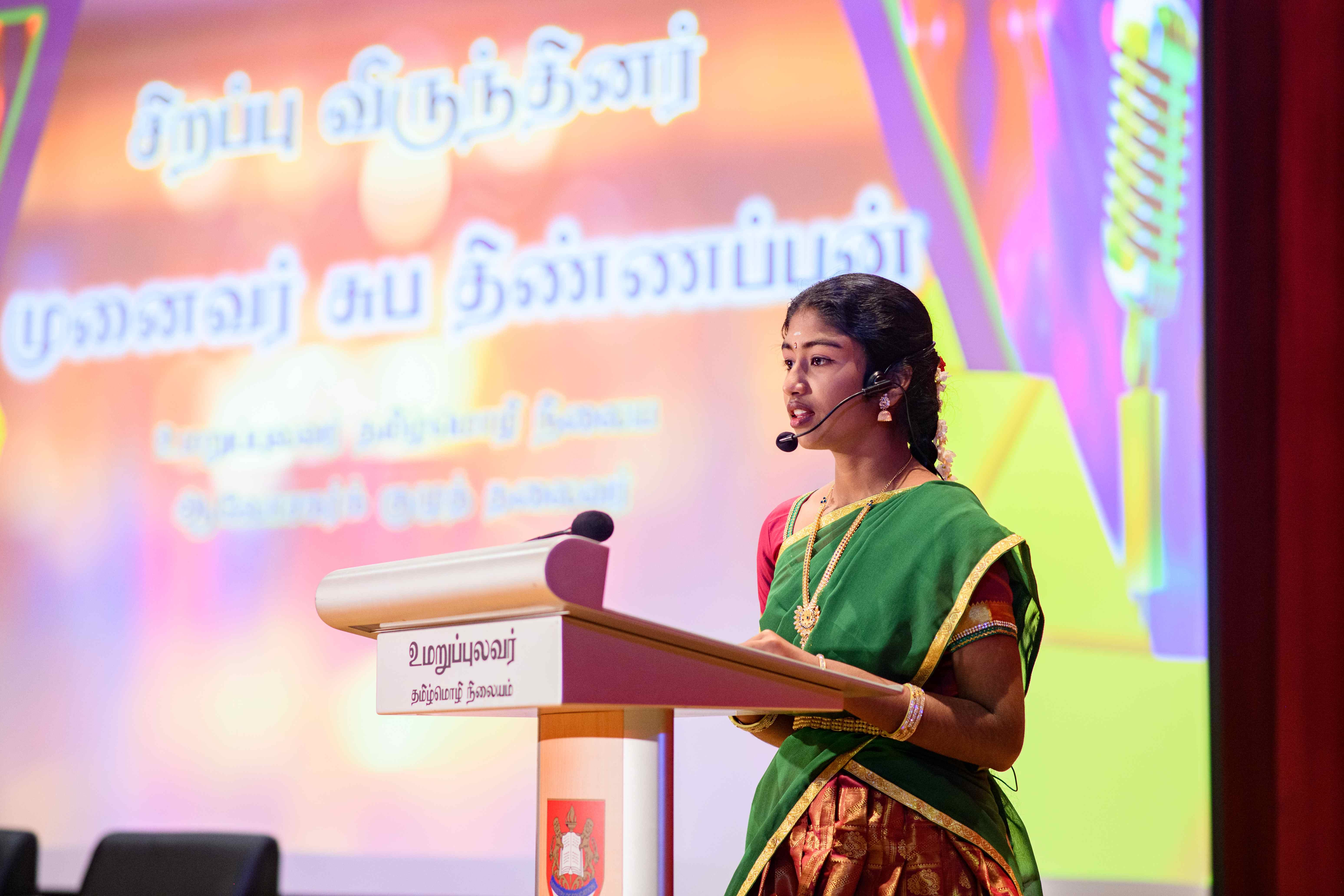 Woman in green sari speaks at podium with headset mic; Tamil text and microphone graphic on screen behind.