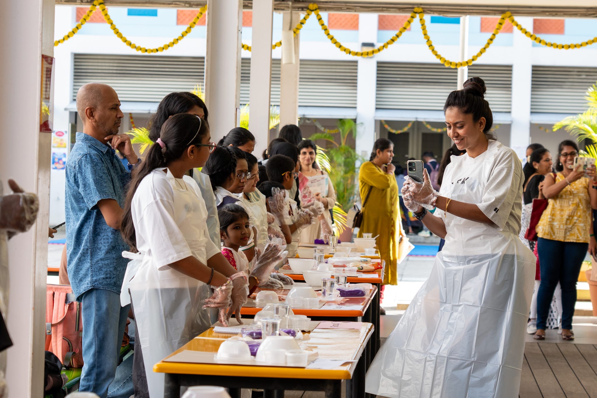 Woman in apron and gloves films people lined at tables with bowls and cups in a decorated school corridor.