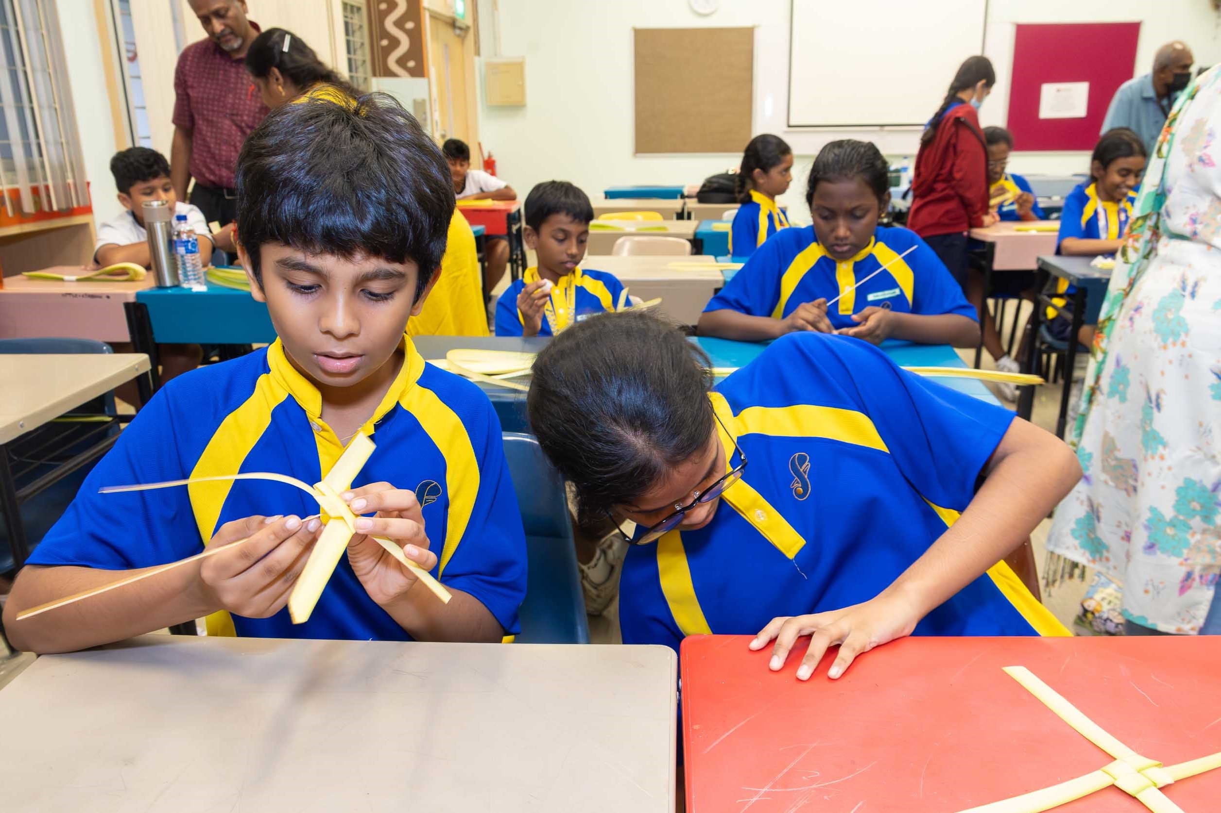 Students in blue-and-yellow uniforms weave palm leaves at desks while others work in a classroom behind them.