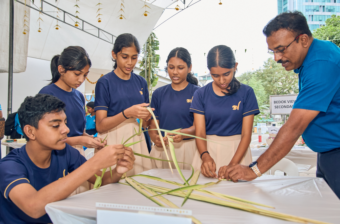 Five students and a man weave long green palm leaves on a table under a canopy.