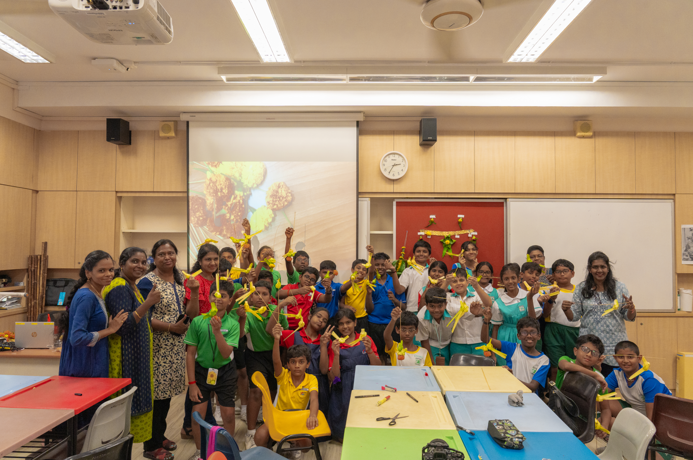 Group of children and adults in a classroom holding yellow paper crafts, with a projector screen behind them.