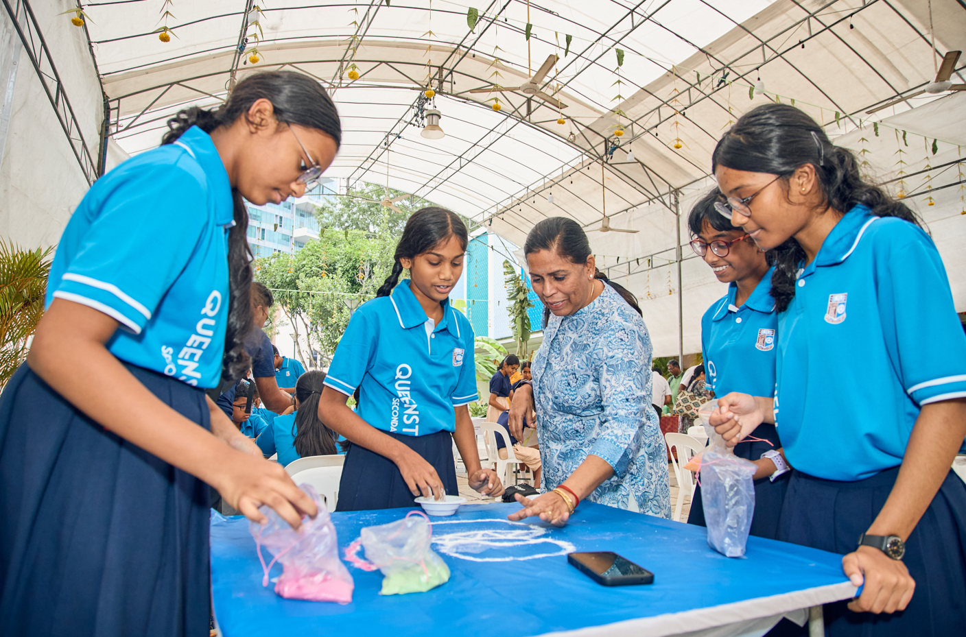 Four schoolgirls and a woman lean over a table, using colored powder in plastic bags under a canopy.