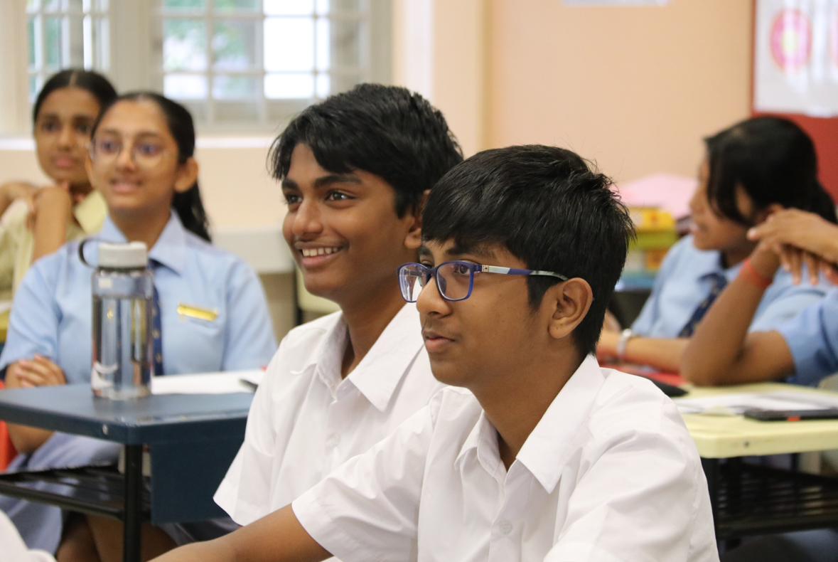 Two boys in white school uniforms sit at desks in a classroom, one wearing blue glasses, smiling.