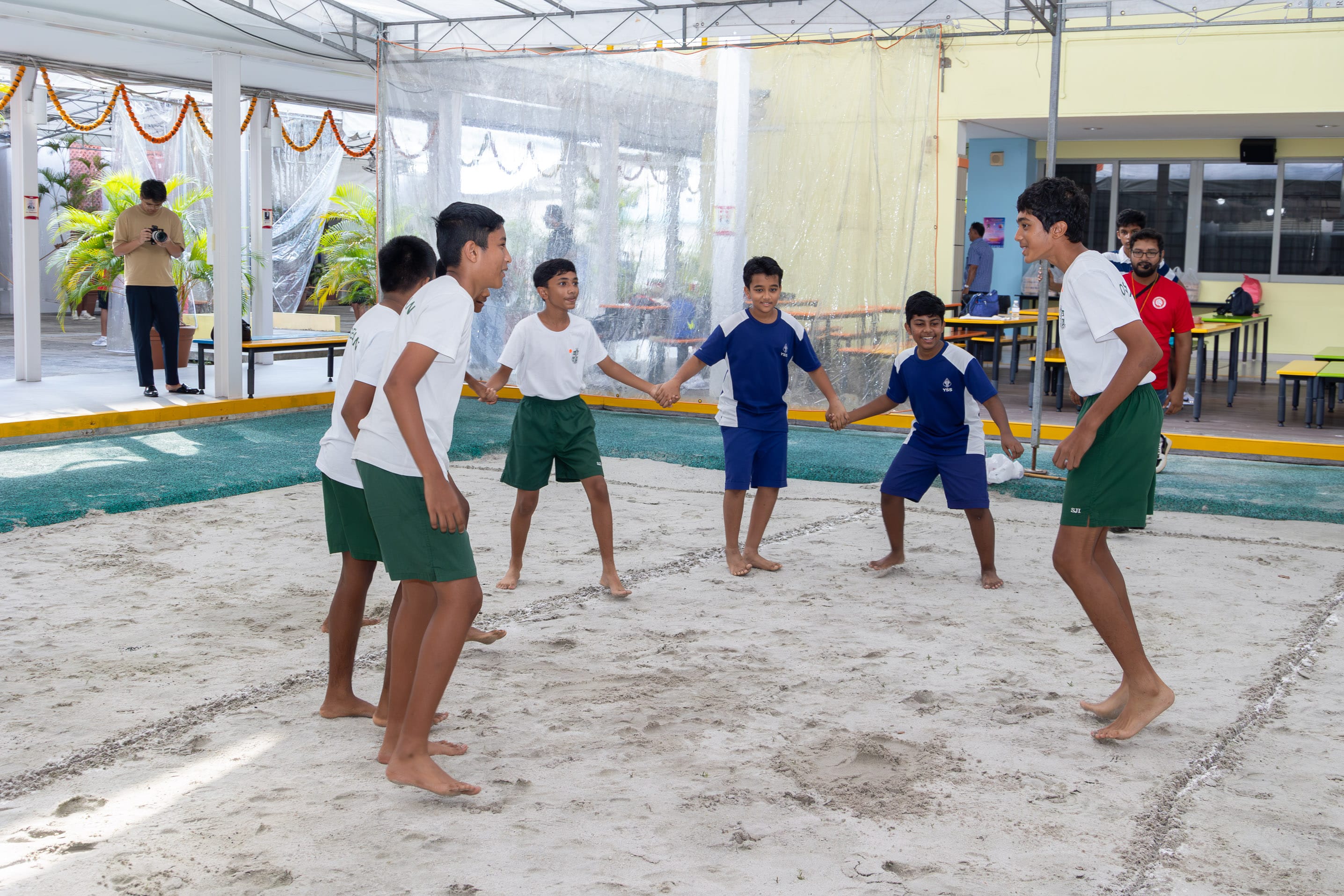 Boys in sports uniforms play a circle game barefoot on a sandy court under a covered school area.