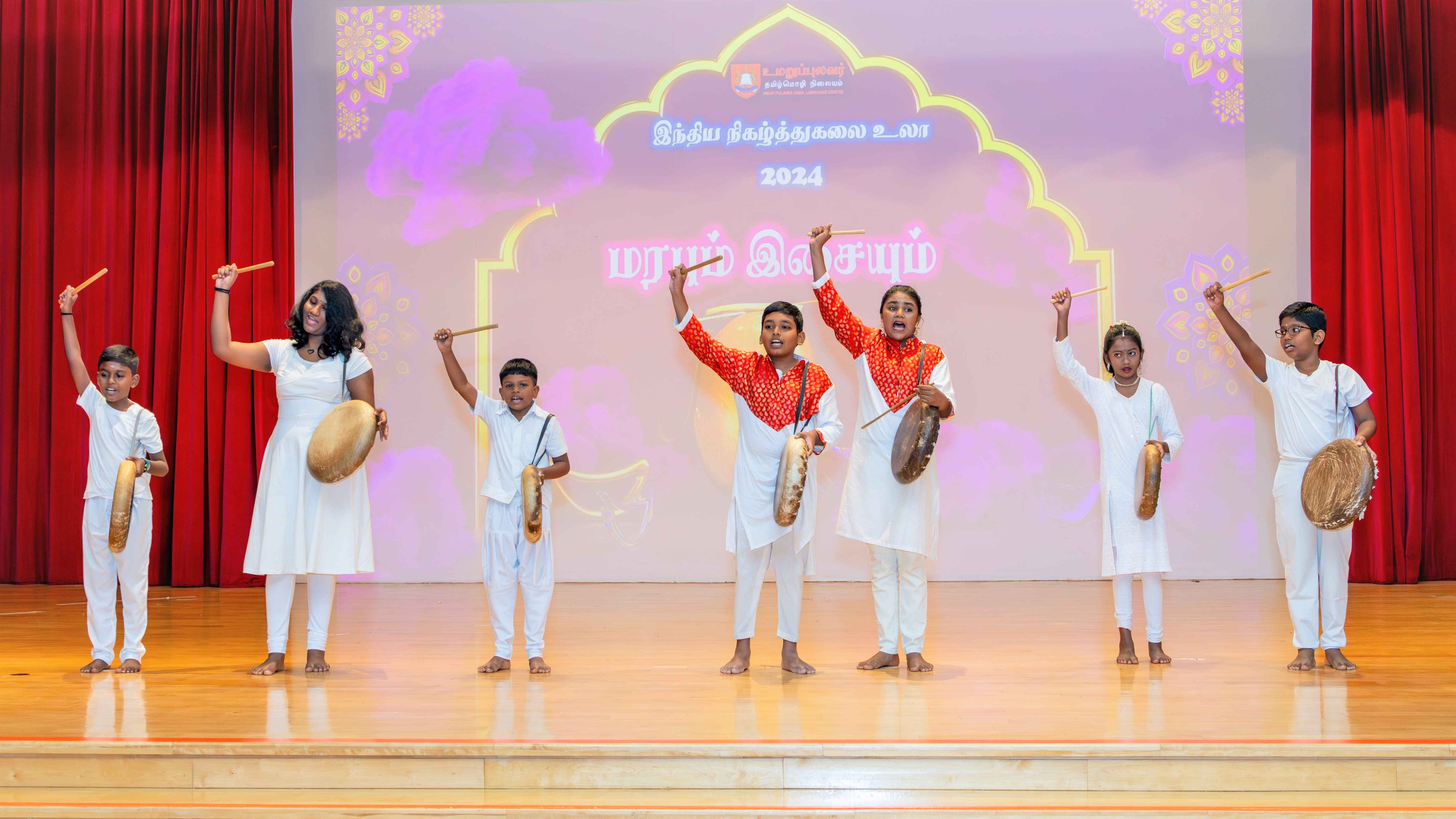 Six students on stage raise sticks and hold drums, wearing mostly white; backdrop shows Tamil text and 2024.