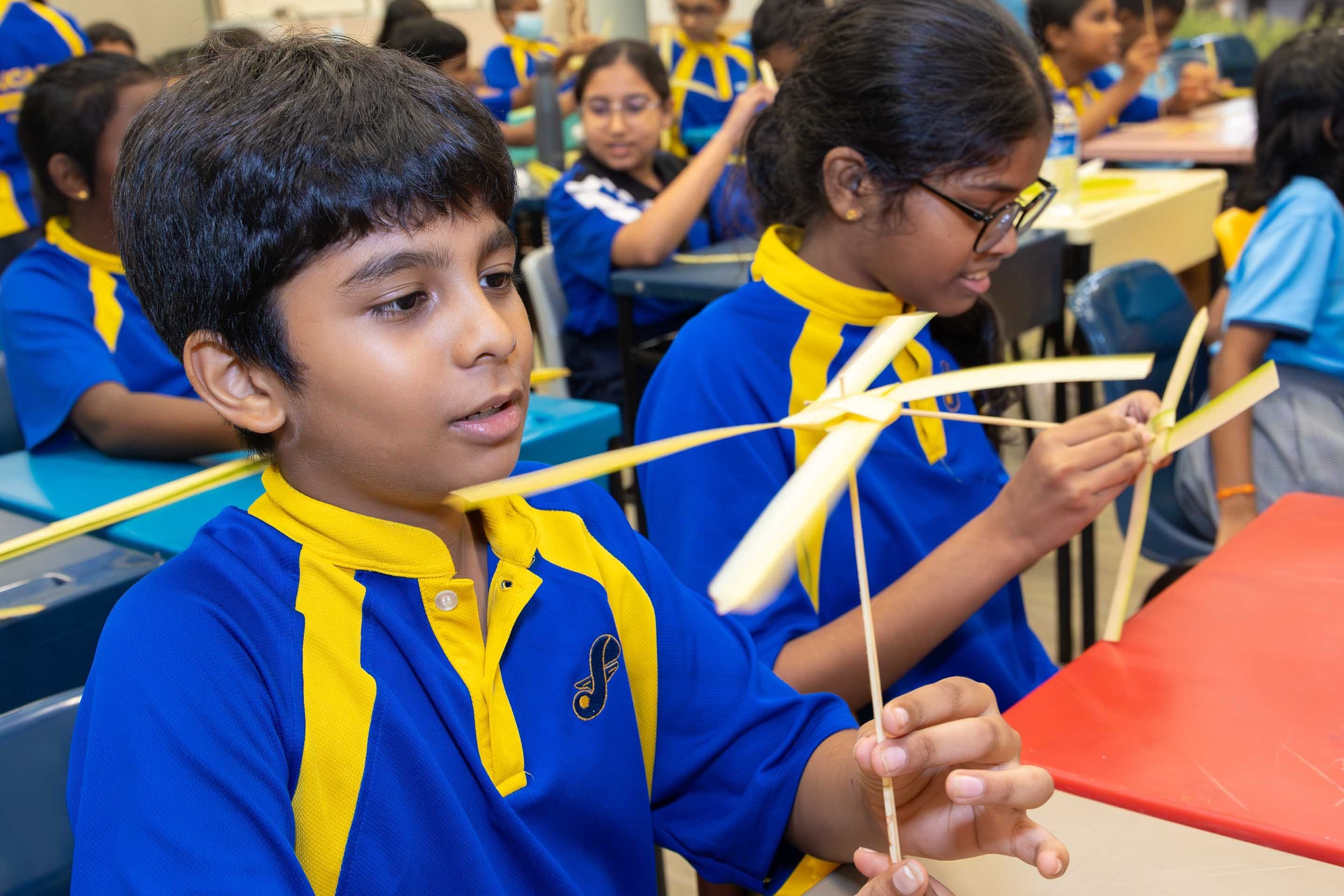 Students in blue-and-yellow uniforms make paper pinwheels at desks in a classroom.