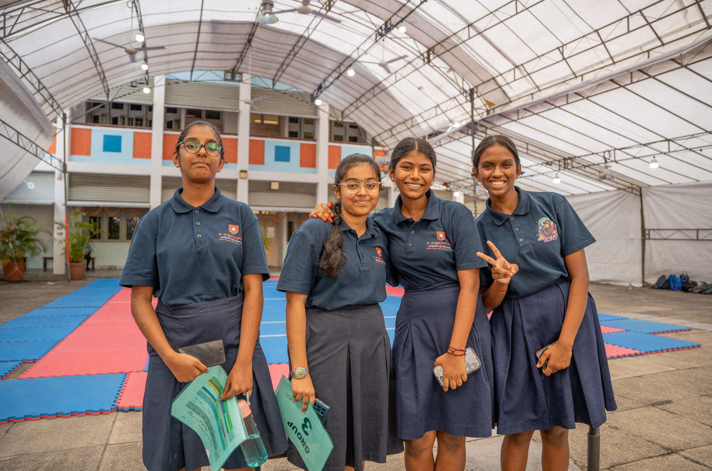 Four schoolgirls in navy uniforms pose under a covered courtyard; one flashes a peace sign.