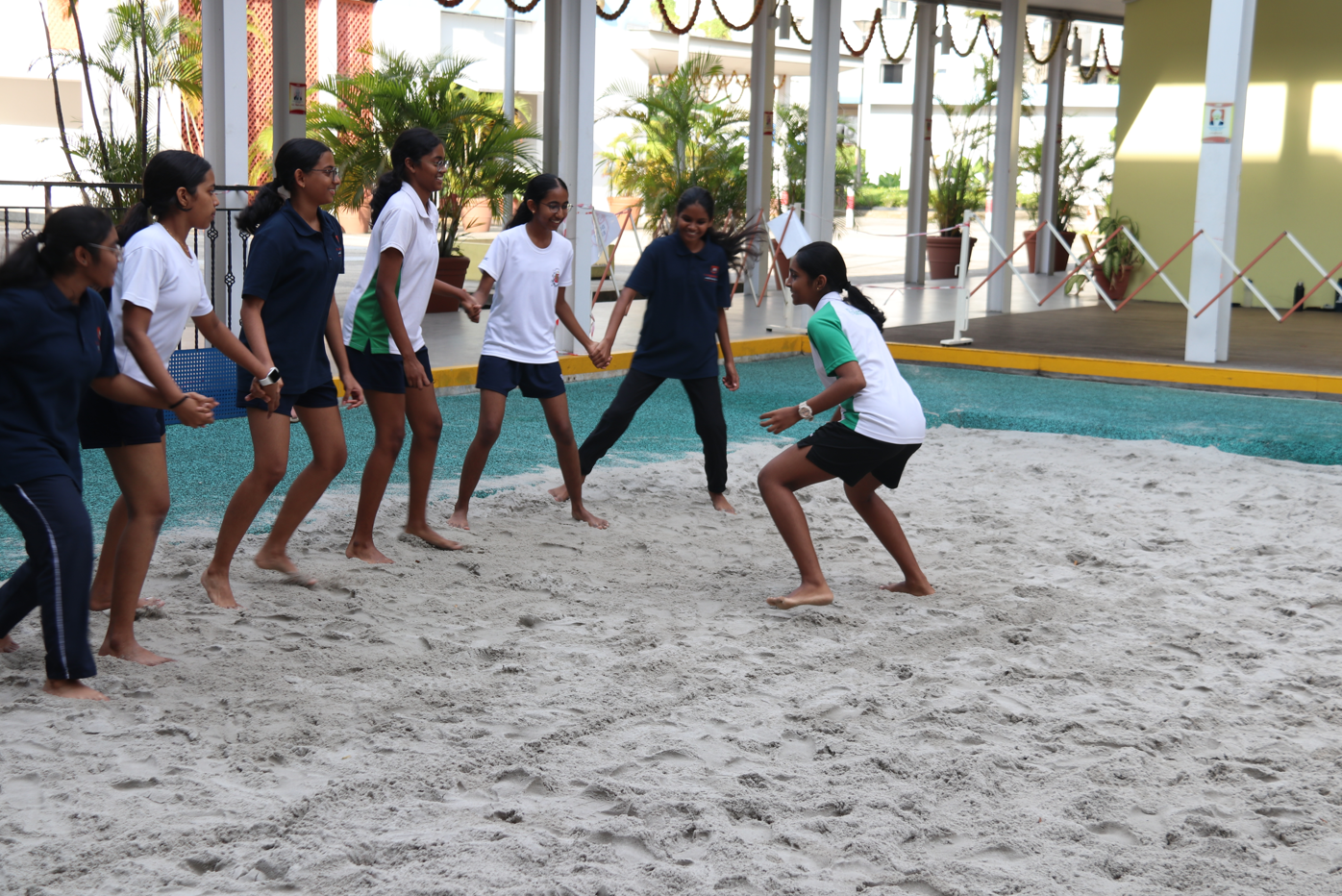 Girls playing a team game barefoot in a sandy court under a covered walkway with plants behind them