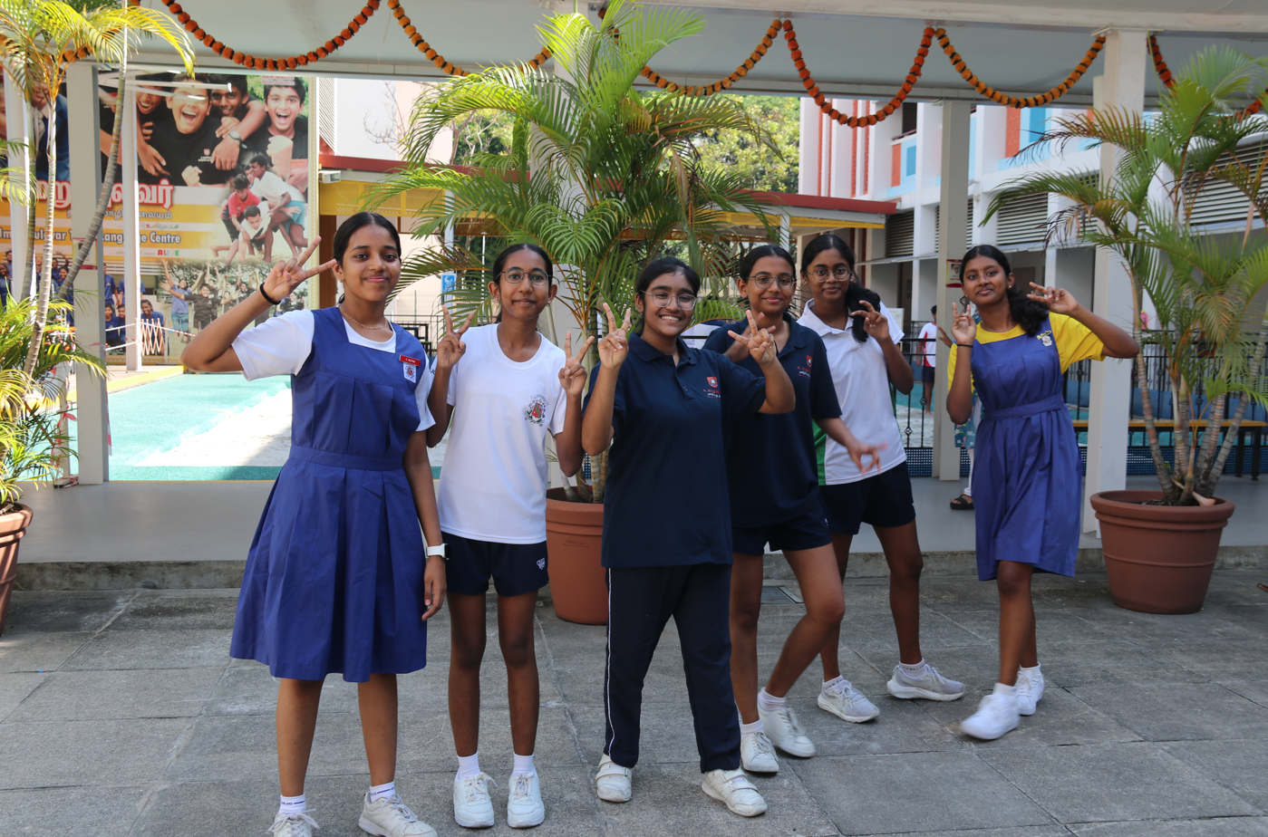 Six schoolgirls pose smiling and making peace signs in a courtyard with potted palms and garlands overhead.
