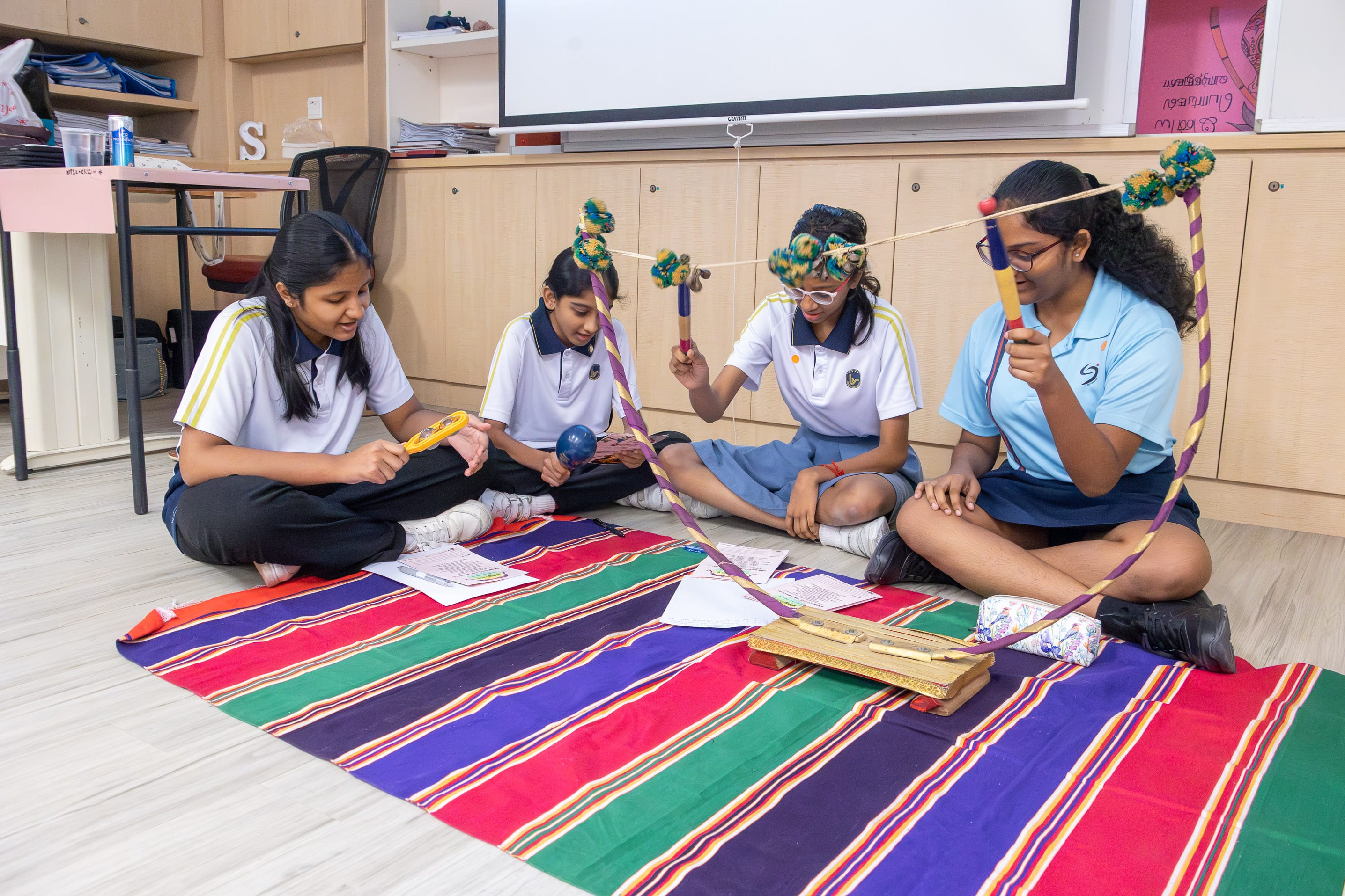 Four students sit on a striped mat, holding sticks linked by string with pom-poms during a class activity.