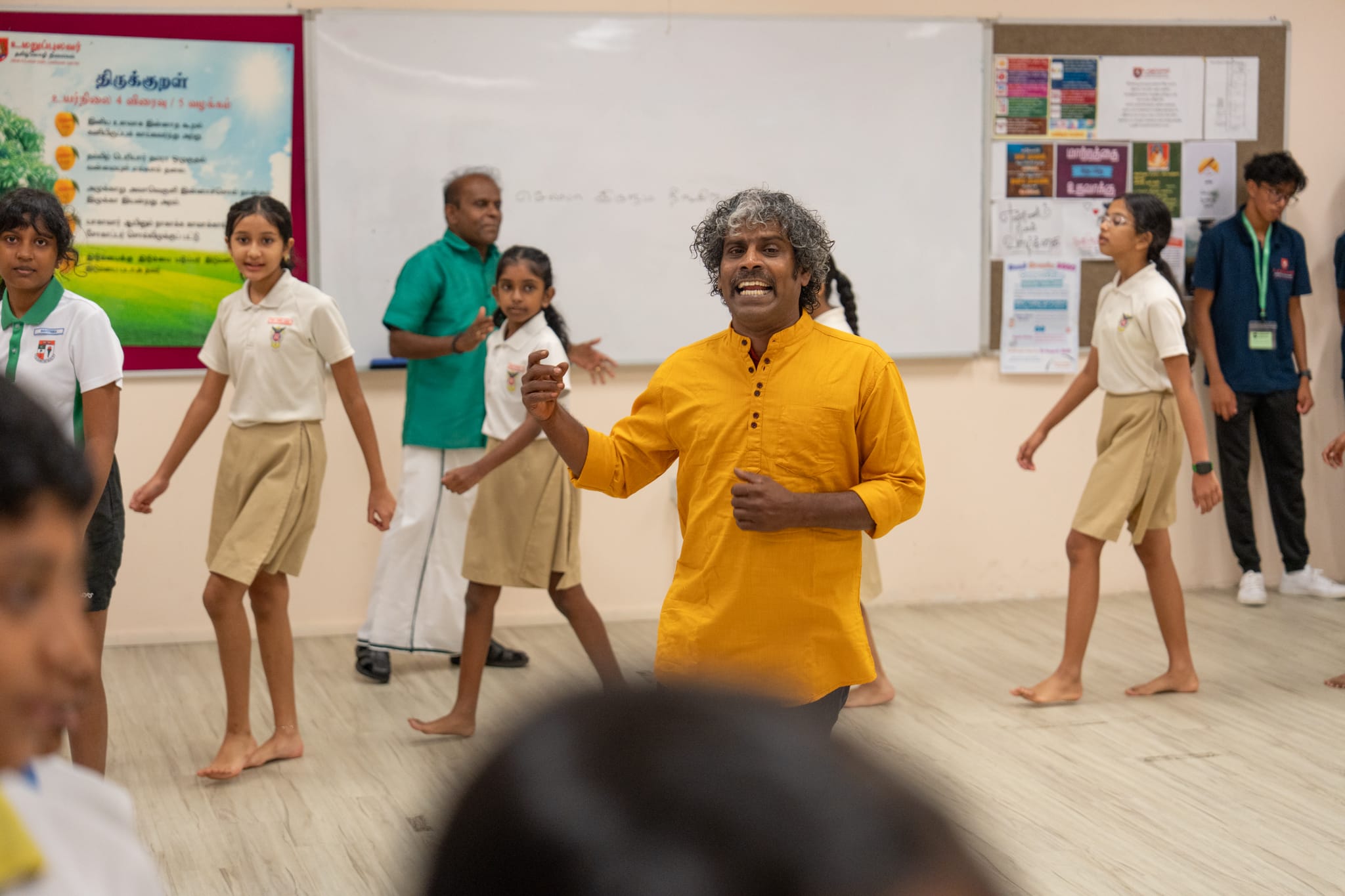 Teacher in yellow leads students in uniform walking barefoot in a classroom with a whiteboard and posters