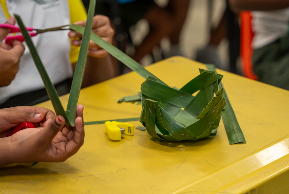 Hands cut green leaves with scissors on a yellow table beside a woven leaf bowl and a small yellow tape measure.