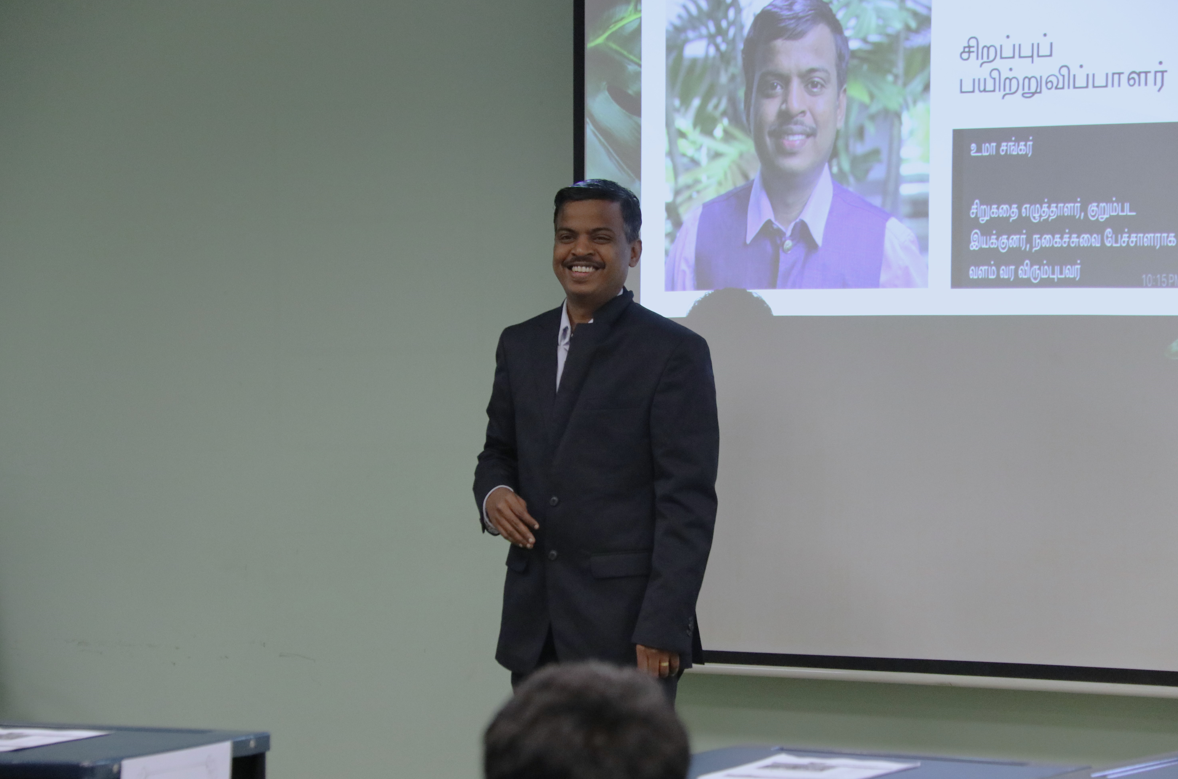Smiling man in a dark suit stands by a projector screen showing his photo and Tamil text.