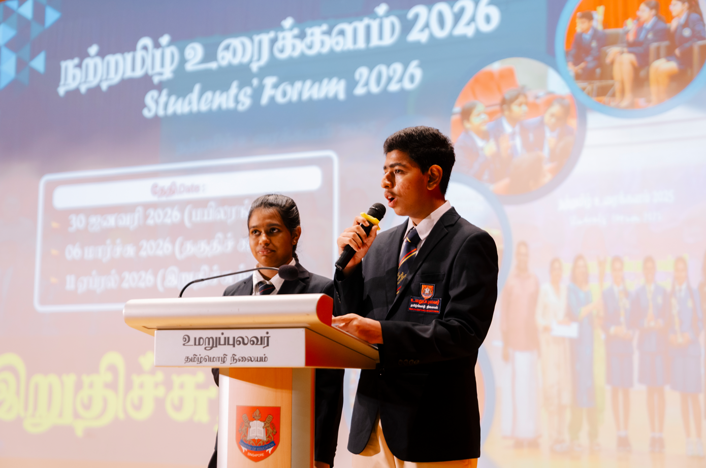Two students in school uniforms speak at a podium on stage before a banner reading Students' Forum 2026.