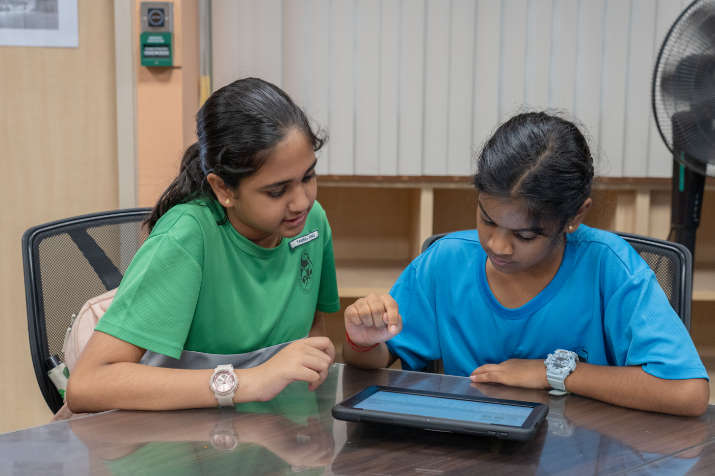 Two girls sit at a table, focusing on a tablet; one points at the screen while the other watches.